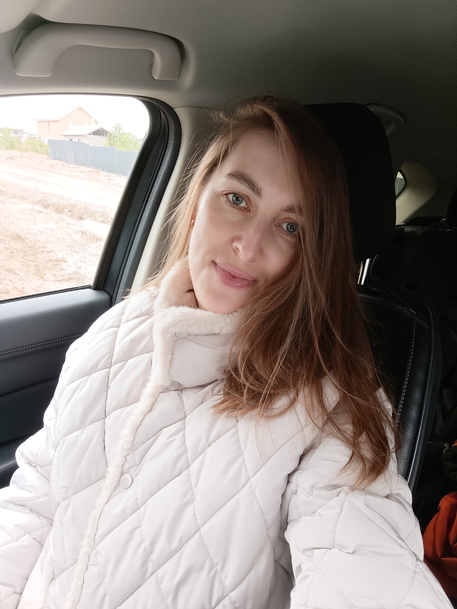 This image captures a woman seated in the passenger side of a car, looking directly at the camera with a calm, subtle smile. She has long, flowing reddish-brown hair and light blue-green eyes. She is wearing a cream or off-white quilted puffer jacket, featuring a soft, fluffy trim along the collar and down the front, secured with white buttons.
Inside the car, elements of the interior are visible, including a black headrest and part of a black leather car seat to her right, and a dark car door panel to her left. A light gray overhead grab handle can be seen against the car's headliner.
Through the car window, an outdoor scene unfolds under what appears to be an overcast or cloudy daytime sky, resulting in soft, diffused lighting. The landscape outside shows dry, unpaved ground, suggesting a dirt road or undeveloped area. In the distance, a light-colored house or building, possibly under construction, is visible behind a fence, with sparse vegetation around it. The woman's warm jacket suggests cool weather. The scene portrays a casual moment during a journey through a rural or developing suburban area, aligning with the context of Chaikovsky, Russia. No text is visible in the media file.