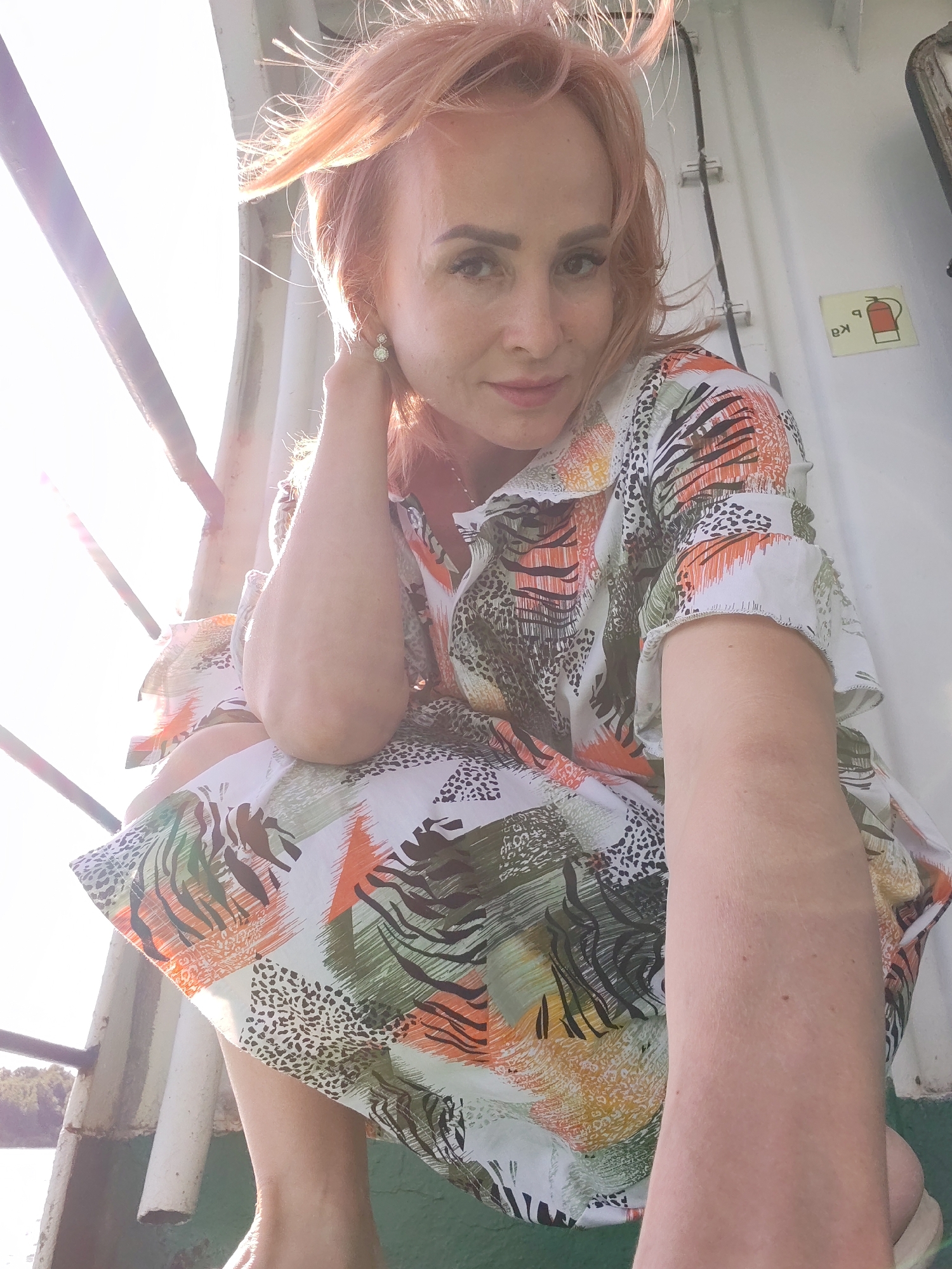 This media file shows a woman with light pink hair, wearing a colorful dress with an abstract print. She is looking at the camera with a slight smile, her left hand touching her neck. The background appears to be outdoors, possibly on a boat or ferry, with a railing and a white structure behind her. Sunlight is coming from the left, creating a lens flare and illuminating her hair and face. A sign with a fire extinguisher symbol is visible on the white structure. The scene suggests a casual, daytime setting with warm weather. The location context provided is Nova, Russia.