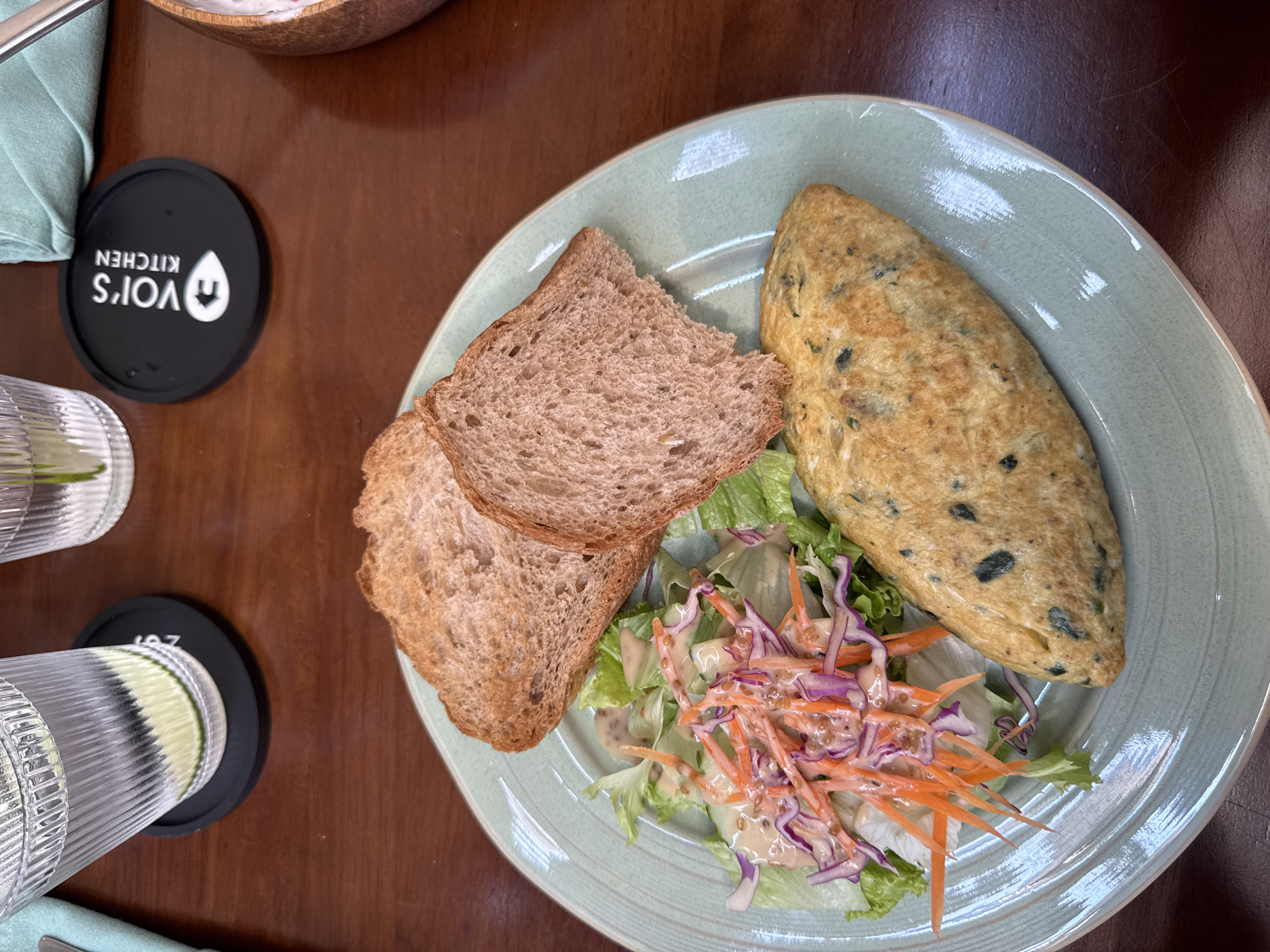 The image depicts a plate of food on a wooden table. The plate contains an omelet, two slices of brown bread, and a salad with lettuce, shredded carrots, and purple cabbage, dressed with a light-colored sauce. To the left of the plate are a glass of water with lemon or lime slices, a black coaster with white text "VOR'S KITCHEN" in reverse, and another black coaster with the letters "AOZ". A wooden bowl is visible in the top center of the frame. A light blue napkin is also partially visible to the left. Given the text "VOR'S KITCHEN" on the coaster, the location is likely Đà Nẵng, Vietnam, where a restaurant with that name exists. The scene suggests a casual dining setting.