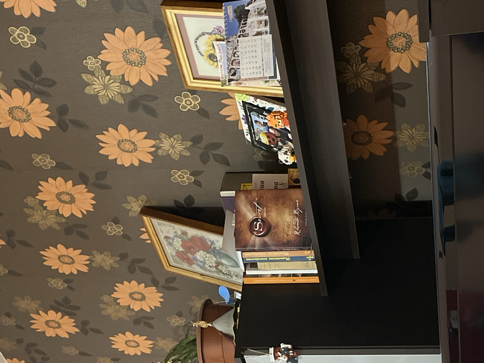 The image displays a close-up of a shelf in a domestic setting, with a dark brown floral wallpaper as the background. The shelf holds several items, including framed pictures, books, and small decorative figures.
On the shelf, from left to right, there is a dark brown cabinet or shelf unit. To its right, there are several books, one of which has a prominent cover featuring a red wax seal and the word "Secret" in gold lettering. Next to these books are two framed pictures. The first frame contains a photograph of people. The second frame has a calendar with images of a landscape and a sunflower basket. There are also some small decorative figures on the shelf, including a panda and an orange tiger.
The wallpaper has a repeating pattern of large orange and gold flowers with dark leaves. The lighting suggests an indoor environment, likely a home. The scene is static, with no visible actions or interactions occurring.
The image does not provide any direct information about the location in the city of Plovdiv, Bulgaria. It is a generic interior scene.