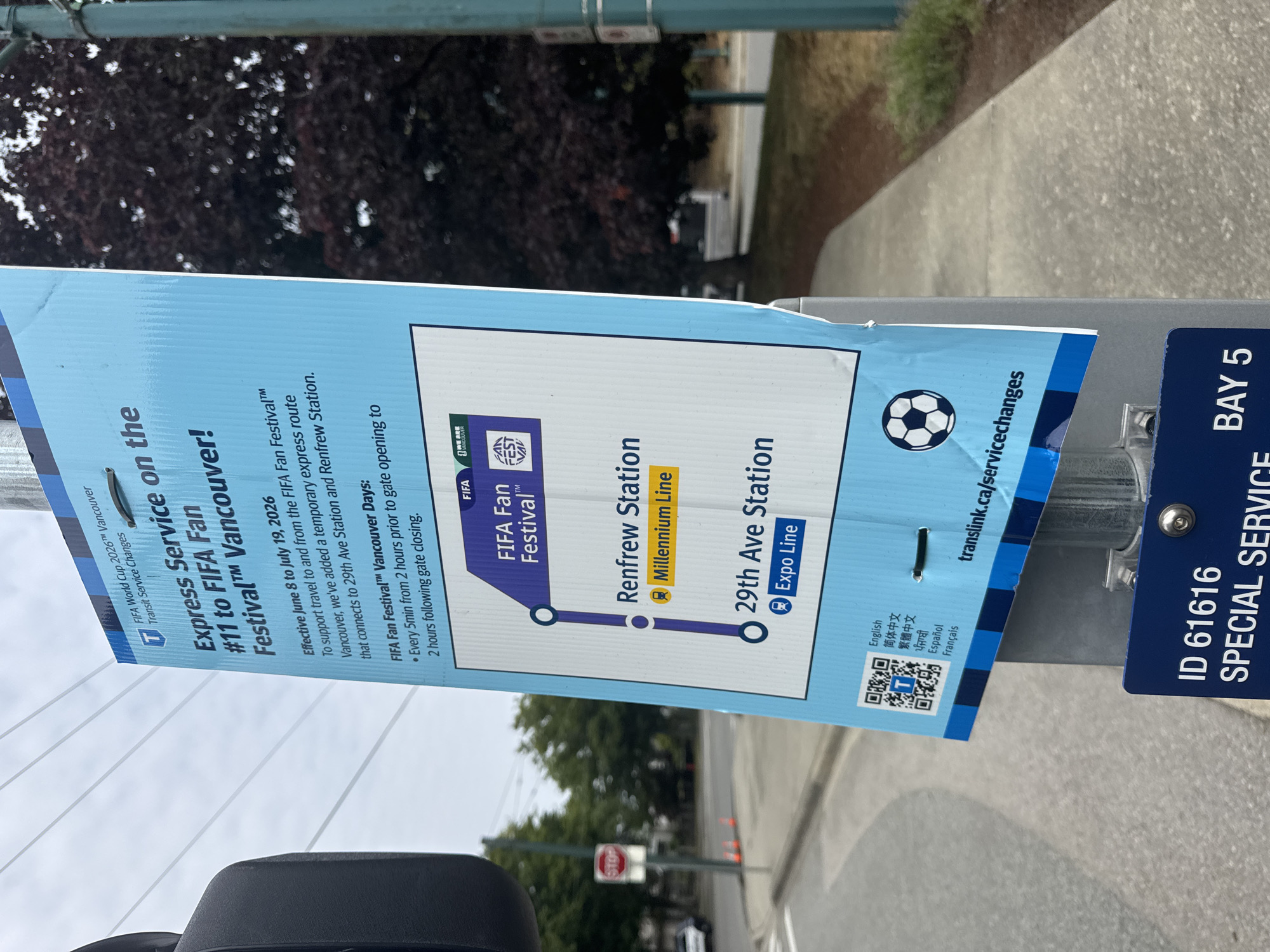 The image shows a blue and white sign on a pole near a street in Vancouver, Canada. The sign announces "Express Service on the #11 to FIFA Fan Festival™ Vancouver!". It details that effective June 8 to July 19, 2026, a temporary express route will connect to 29th Ave Station and Renfrew Station. The route diagram shows the FIFA Fan Festival as the destination, with connections to Renfrew Station (Millennium Line) and 29th Ave Station (Expo Line). The sign also includes QR codes, multilingual text, and a soccer ball graphic. In the background, a traffic light, a stop sign, orange cones, trees, and a sidewalk are visible. A dark blue sign with "ID 61616 SPECIAL SERVICE BAY 5" is attached to the same pole below the main announcement. The overall scene appears to be an outdoor transit stop in a city environment.