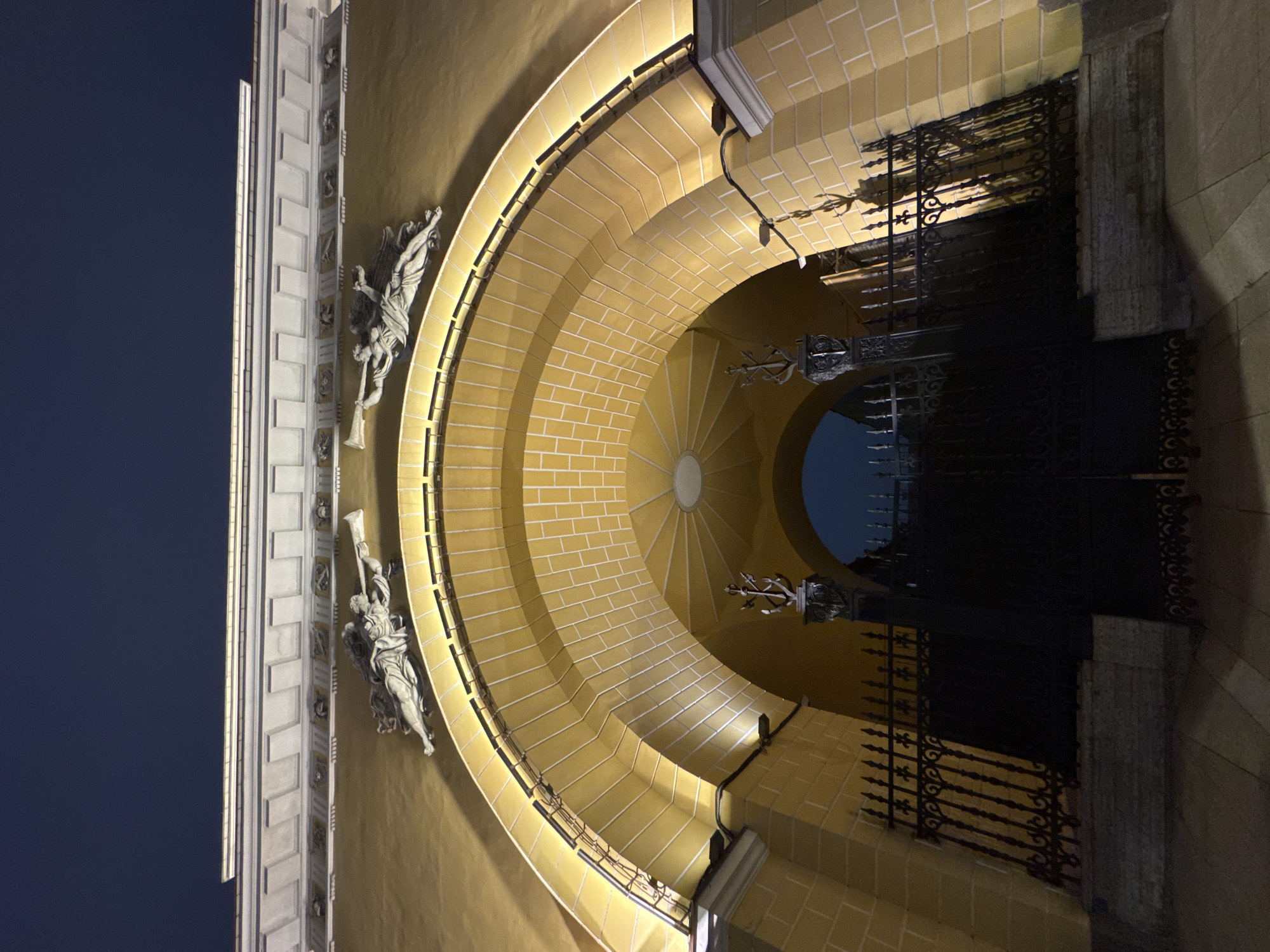 The image depicts an illuminated entrance to a building at night. Two angel statues flank a golden-yellow, brick-patterned archway leading to a black wrought iron gate. The sky is visible through the gate. A building facade is to the left. The location is Saint Petersburg, Russia.