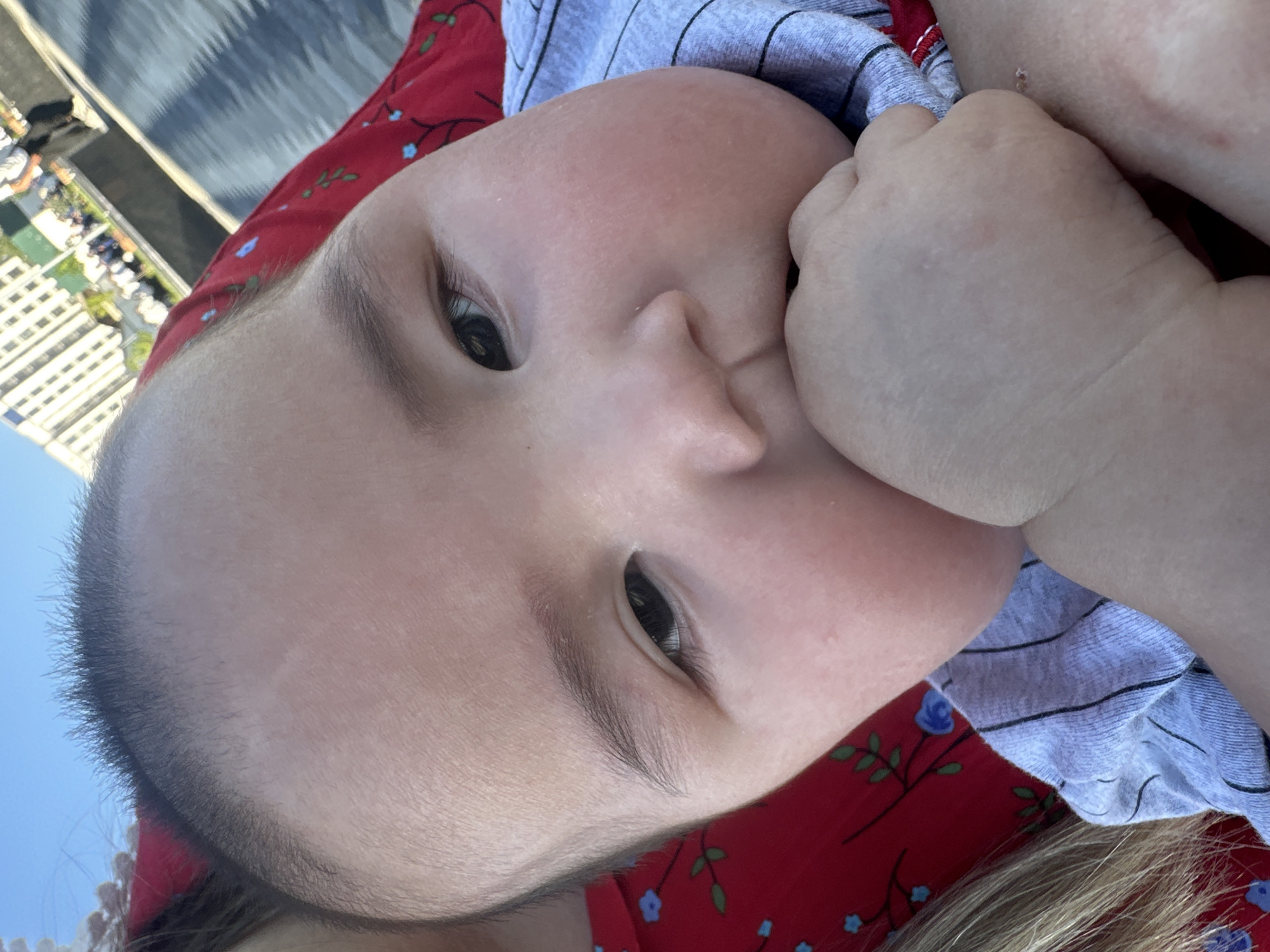 A close-up, head-and-shoulders shot captures a baby's face, with the majority of the frame occupied by the child's head and a portion of their body. The baby's right hand is brought up to their mouth, with fingers partially inserted. The baby is wearing a gray, striped shirt. Behind the baby, a red fabric with a floral print is visible, along with a strand of blonde hair. The background is slightly out of focus, showing a blue sky and buildings that appear to be in an urban setting, possibly Manas, Kyrgyzstan. The lighting suggests it is daytime, with the sun casting shadows.
