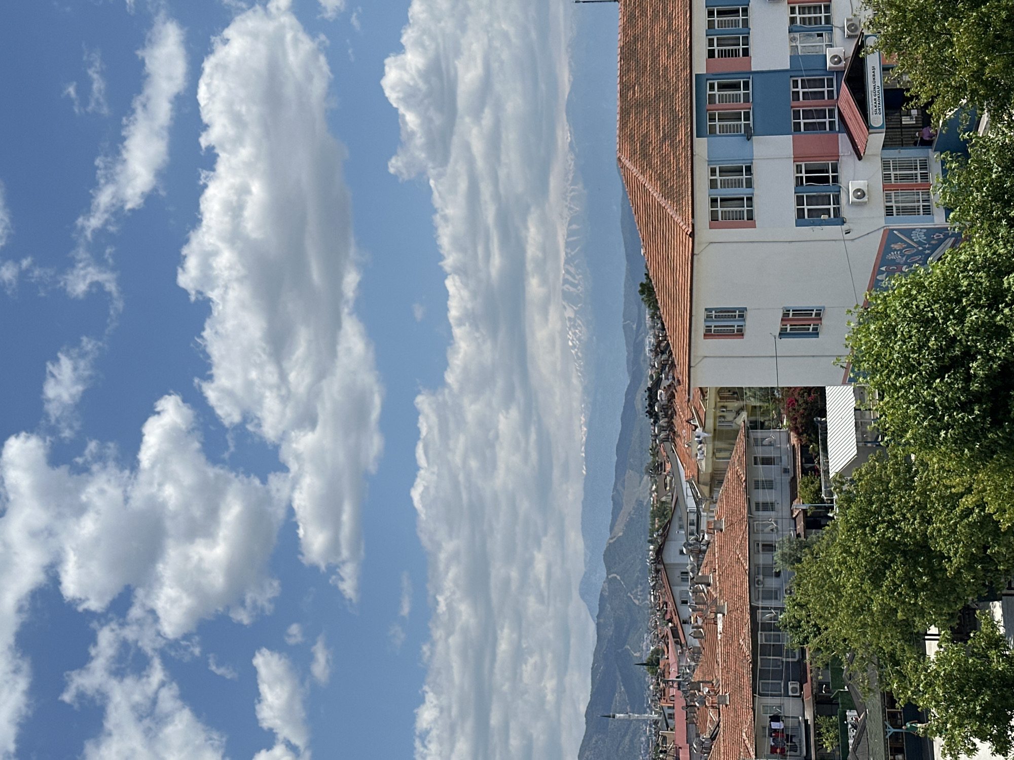 The image shows a cityscape in Fethiye, Turkey, under a partly cloudy sky. The view encompasses urban architecture and distant mountains. Predominantly, buildings with red-tiled roofs line the scene, intermingled with multi-story structures. One taller, white building is partly visible, featuring blue and pink sections on its facade and several air conditioning units mounted on the exterior. A school is identified by the signage “SILKAR GÜNLÜKLU BASI ORTAOKULU". Trees are visible on the right side of the image. In the distance, there is a mountain range. The sky is filled with cumulus clouds and a clear blue background.