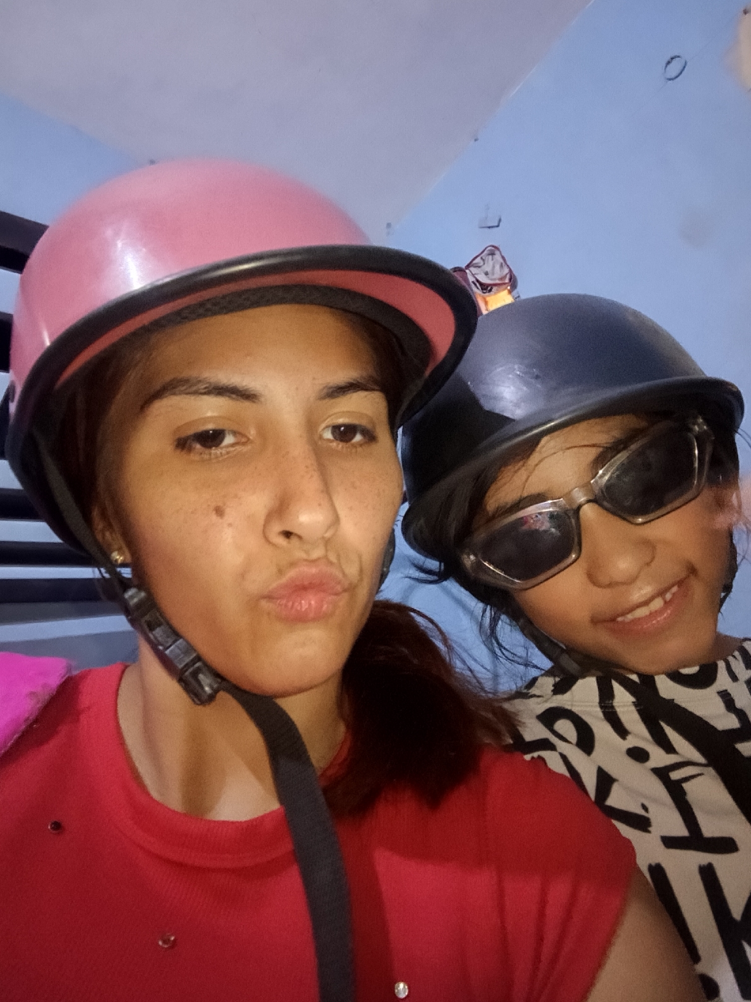 Two individuals are captured in a close-up shot, both wearing helmets. The individual on the left is an adult female with fair skin and freckles, donning a pink helmet and a red shirt. She is making a kissing face towards the camera. The individual on the right appears to be a child with dark sunglasses and a black helmet. They are wearing a white shirt with black lettering. The background suggests an interior setting with light blue walls and a dark horizontal element, possibly part of a structure or furniture. The exact location is Juan Griego, Venezuela.