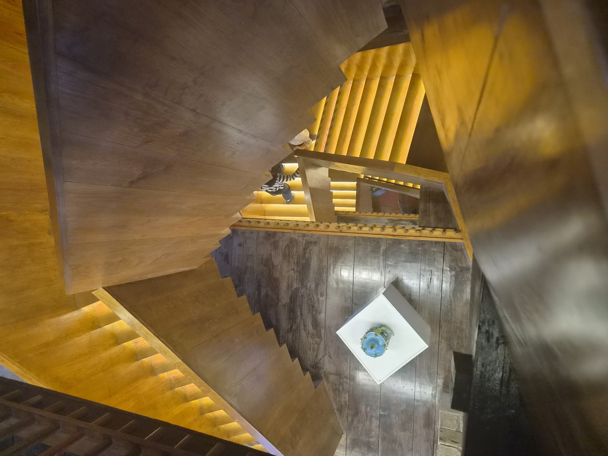 The image shows an interior view of a modern wooden staircase with warm yellow lighting. The perspective is from above, looking down the winding stairs. On the wooden floor below, there is a white cube-shaped pedestal with a small, blue, decorative object on top. In the background, a person wearing a striped shirt and dark pants is partially visible on the stairs, appearing to be moving upwards. The architecture suggests a contemporary building, possibly a public space or a unique residence. The overall scene is well-lit, emphasizing the natural wood textures and the geometric design of the staircase. This location is not identifiable as being in Barcelos, Portugal from the image alone.