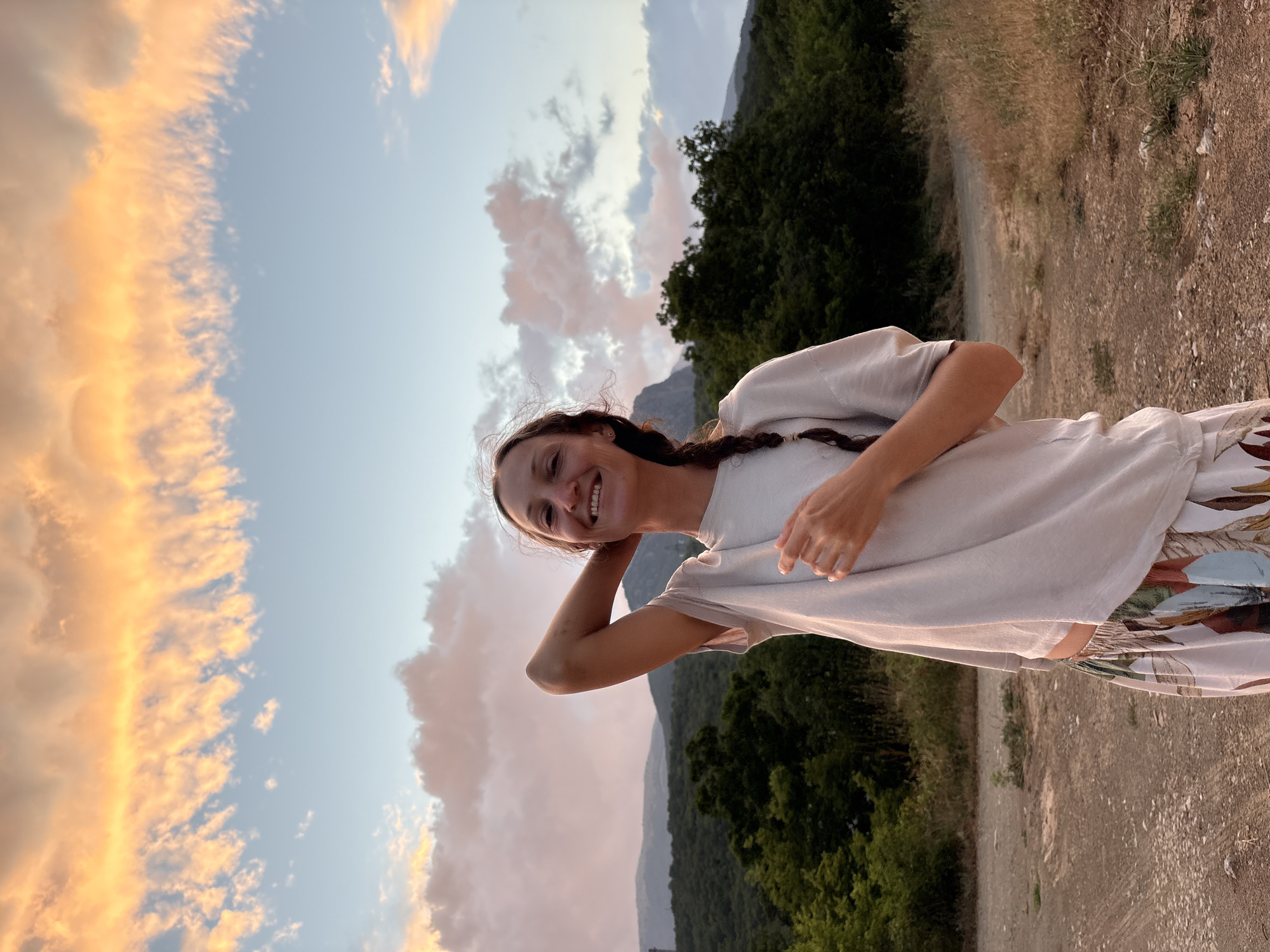 A full-body, outdoor, waist-up medium shot features a smiling young woman with long, dark hair braided down her back, posing with her right arm behind her head. She is wearing a light grey t-shirt and patterned pants with large green and white floral designs. The woman stands on a dirt path with dry grass and brush to her right and a line of dark green trees and rolling hills behind her. The sky above is a soft blue, with wispy clouds illuminated by a warm, orange glow, suggesting it is either sunrise or sunset. The location is the village of Malyi Mayak, Ukraine.