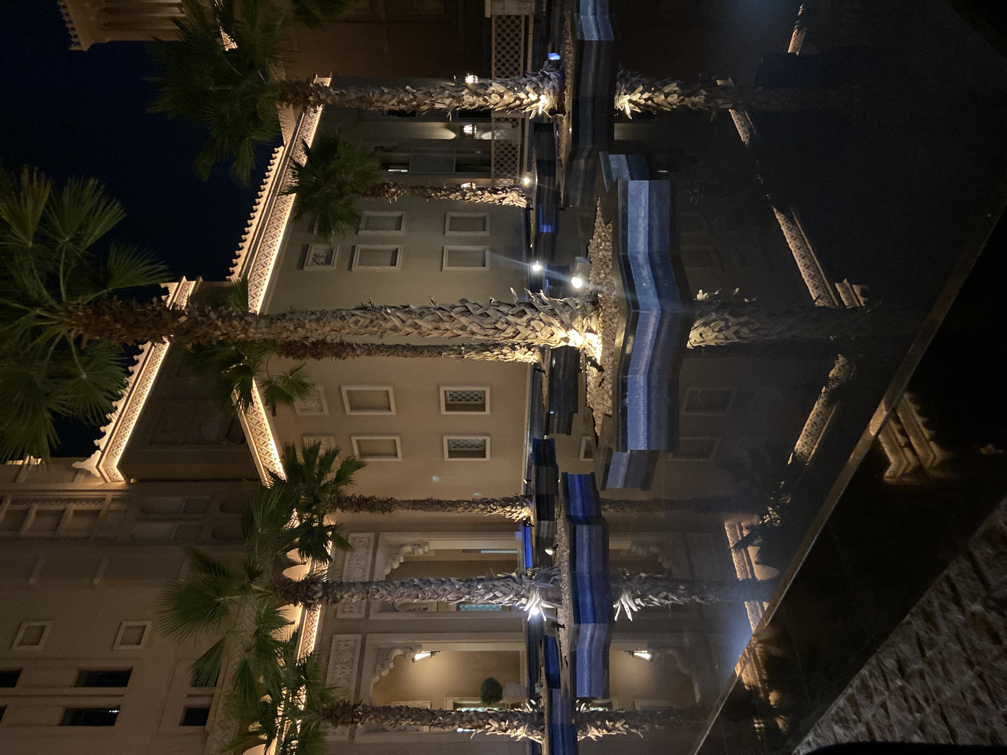 The image depicts a nighttime scene, seemingly in Dubai, United Arab Emirates, featuring a building with ornate architectural details illuminated by soft lighting. Rows of palm trees line the building, their trunks and fronds clearly visible. The image is partially reflected in a dark, glossy surface, possibly water, creating a mirrored effect that distorts the scene. The building appears to have multiple floors with windows and decorative archways. The lighting emphasizes the building's design, particularly the intricate patterns along the roofline and facade. The overall impression is one of a tranquil and luxurious environment, typical of Dubai's urban landscape.