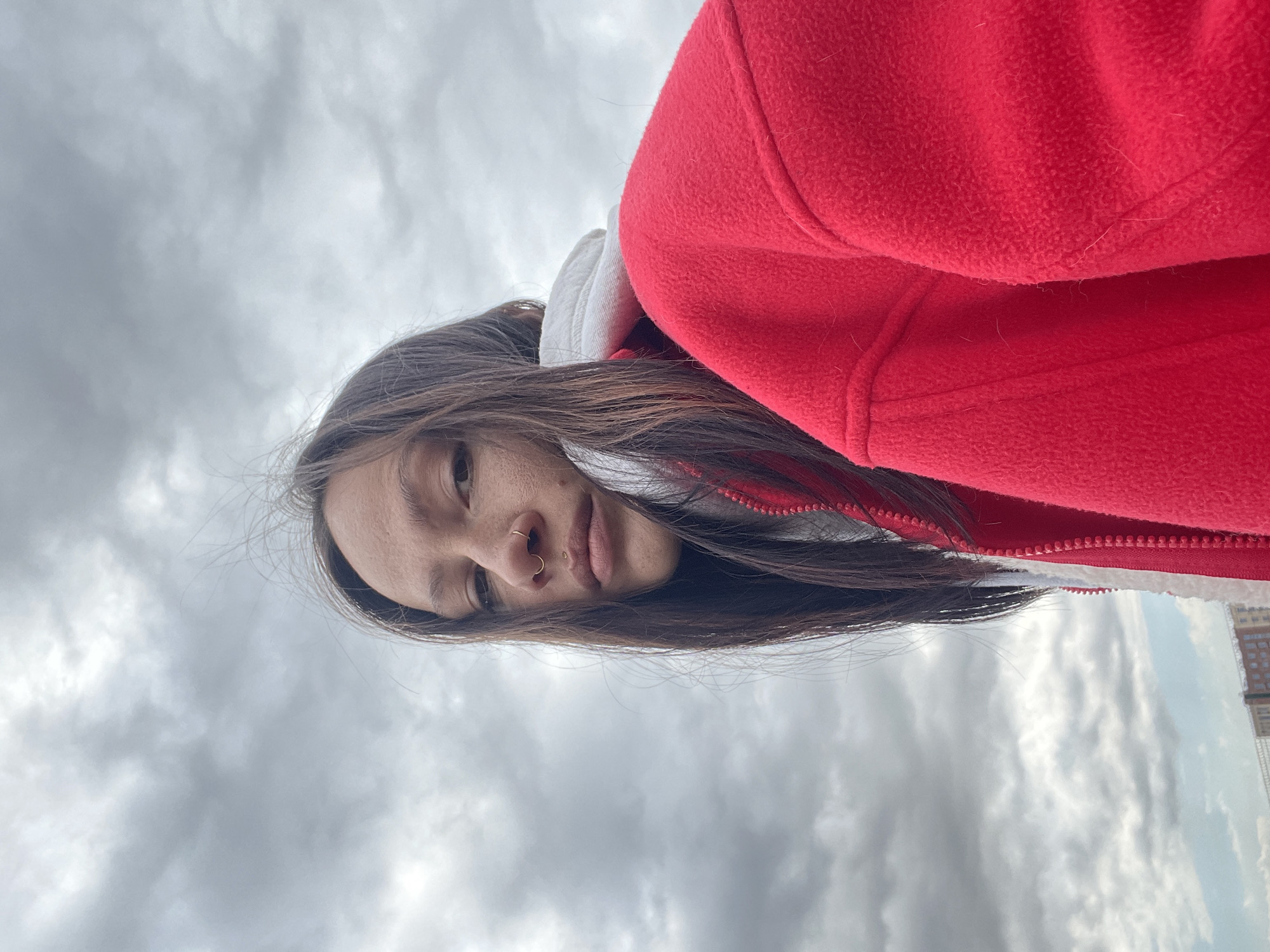 A close-up, eye-level shot features a person, likely a young woman, outdoors against an overcast sky. The person is looking directly towards the camera with a neutral expression. She has long, dark hair that falls over her shoulders and part of her face. Notable facial features include a golden hoop nose ring in her right nostril and a small, gold-toned stud piercing above the center of her upper lip. She is wearing a vibrant red fleece jacket with a visible white inner lining or hood. The texture of the fleece fabric is apparent, showing small fibers. The sky behind her is filled with various shades of grey clouds, indicating an overcast or cloudy day. On the far right side of the image, barely visible on the horizon, appears to be the top section of a building, suggesting an urban or suburban setting. No text is visible in the image.