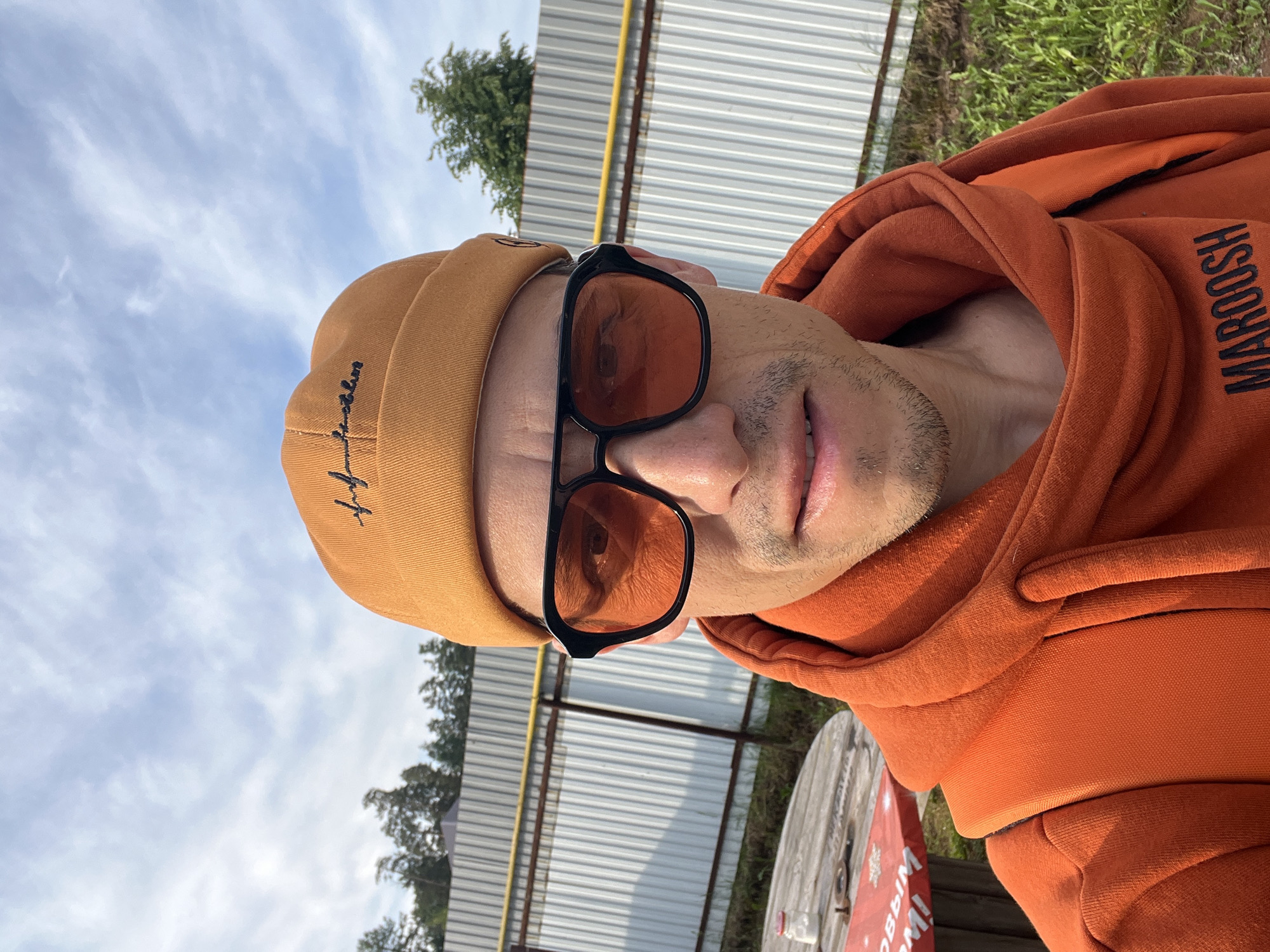 A close-up portrait shows an individual, presumably male, facing the camera. The image is rotated 90 degrees counter-clockwise, making the person's head appear tilted to their left. The subject wears an orange beanie with dark, stylized script embroidery on the side, and dark-framed sunglasses with orange-tinted lenses. They also have an orange hooded sweatshirt (hoodie) with the word "MAROOSH" printed in black capital letters vertically on the right arm/shoulder. The person has light stubble on their chin and upper lip.
The background indicates an outdoor setting during bright daylight, with a clear blue sky and some white clouds. Behind the person, there's a light-colored corrugated metal fence or wall, with a yellow pipe running horizontally across the upper portion. Green foliage, likely trees or bushes, is visible peeking above and behind the fence. In the bottom right corner of the frame, a large wooden spool or reel, possibly for cables, is partially visible. This spool has a red and white sign attached, with partial Cyrillic text visible, suggesting words like "С НОВЫМ И" (S Novym I), potentially part of a greeting like "Happy New Year." The overall location context is Chaikovsky, Russia, suggesting an urban or semi-industrial outdoor environment.