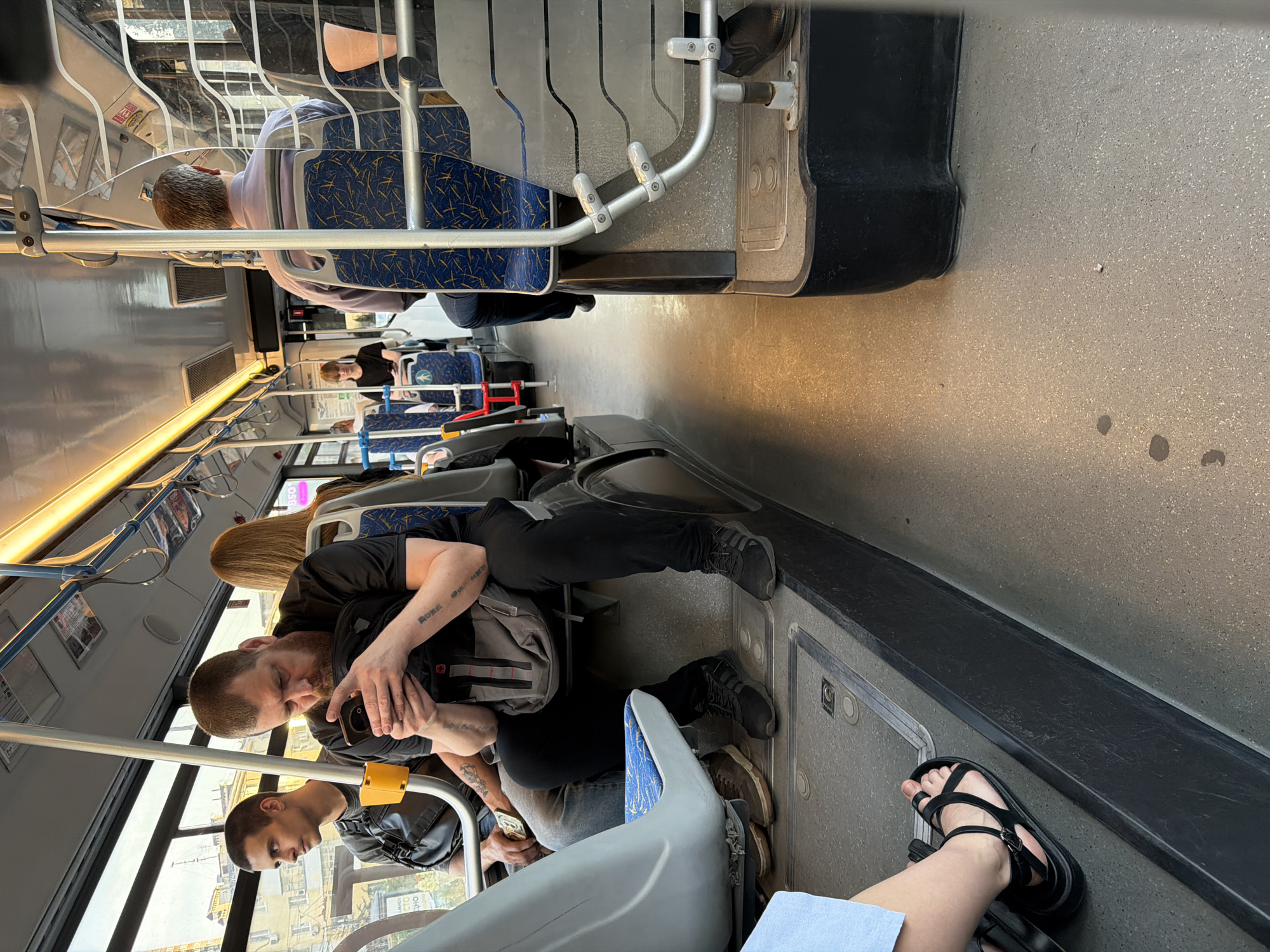 The image depicts the interior of a public bus in Rostov-on-Don, Russia. The perspective is from inside the bus, looking forward and to the right. Several passengers are visible. In the foreground left, a person wearing a light blue garment and black sandals is partially visible. To their right, a man with a beard and tattoos on his arms is seated, looking down at his mobile phone and holding a grey bag. To his left, another man with short hair is looking out the window. Further down the aisle, more passengers are seated, including a woman with blonde hair. The bus interior features grey and blue seats, metal support poles, and windows showing buildings outside. The floor is a grey, speckled material, and the lighting appears to be artificial, coming from overhead fluorescent lights. The overall scene suggests a typical commute on a bus.