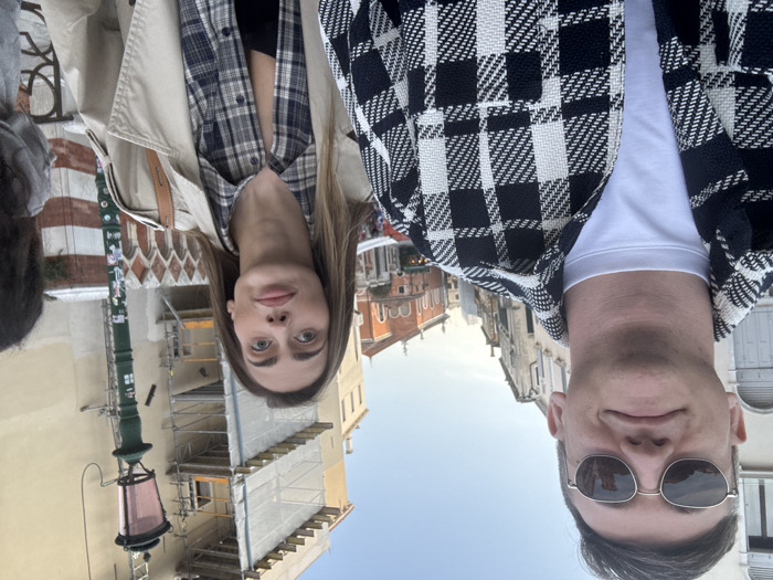 A daytime, eye-level photograph captures three individuals in Venice, Italy. In the left foreground, a male with short dark hair, wearing sunglasses, a white crew-neck t-shirt, and a black-and-white plaid jacket, smiles subtly while looking towards the camera. To his right and slightly behind him, a female with long light brown hair wears a beige trench coat over a blue-and-white plaid button-up shirt and a dark tank top; she stares forward with wide eyes and slightly pursed lips. A third individual, partially obscured by the female's right shoulder, is visible on the far right. The subjects are positioned on what appears to be a bridge or elevated pathway. The background features characteristic Venetian architecture: a decorative barrier or railing with white Gothic-style arch motifs is immediately to the right of the female. Adjacent to this is a tall, dark green lamppost topped with a pinkish-red glass lantern. Behind these elements, a multi-story building is seen undergoing construction, enveloped in scaffolding and white protective netting. Further in the mid-distance, a building with a reddish roof and what appears to be a small dome or cupola is visible, flanked by other traditional, multi-story Venetian buildings. The sky above is clear and light blue.