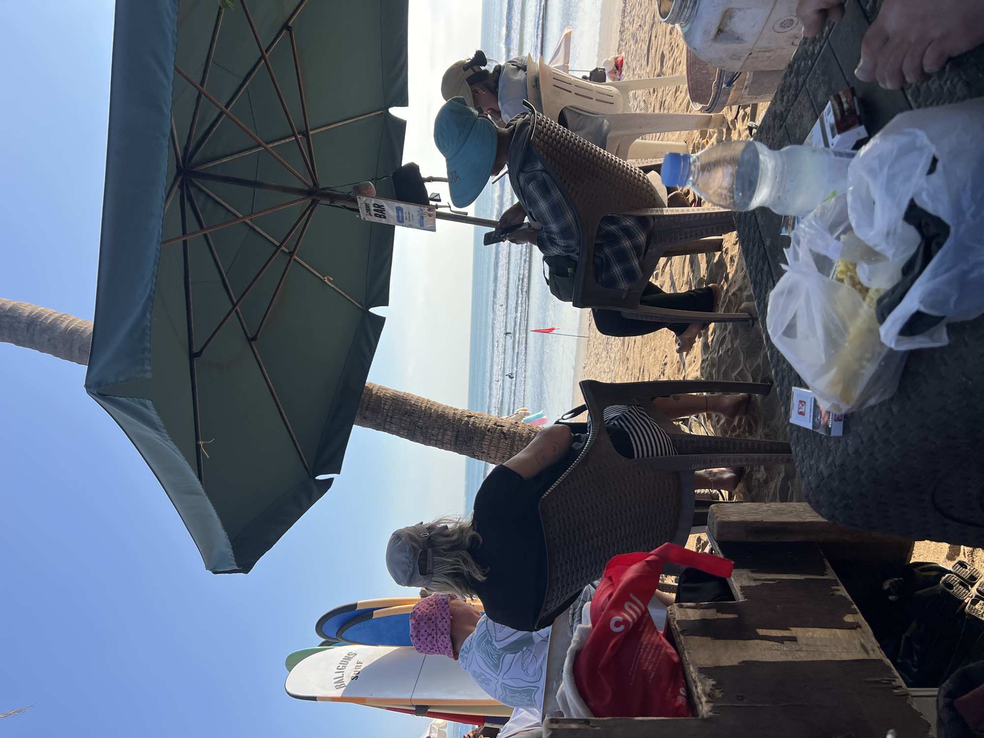 The image depicts a beach scene in Kuta, Indonesia, under a clear blue sky. In the foreground, a rustic wooden structure is visible, laden with a red bag and other items. To the left, several surfboards are propped up, one with "BALIGUNS SURF" written on it. Three individuals are seated under a large, dark green umbrella. The person on the left wears a pink bucket hat and a patterned shirt. The person in the center, facing away from the camera, has long blonde hair and wears a grey cap and a black t-shirt. The person on the right, also facing away, wears a blue wide-brimmed hat and a checkered shirt, and is looking at their phone. In the background, the ocean stretches to the horizon with gentle waves and a few surfers. A red flag is planted on the sand. Further back, other beachgoers and seating arrangements are visible. On a table in the foreground, a pack of cigarettes, a plastic bag with food, and a water bottle are present. The overall atmosphere suggests a relaxed beachside setting, likely a bar or cafe.
