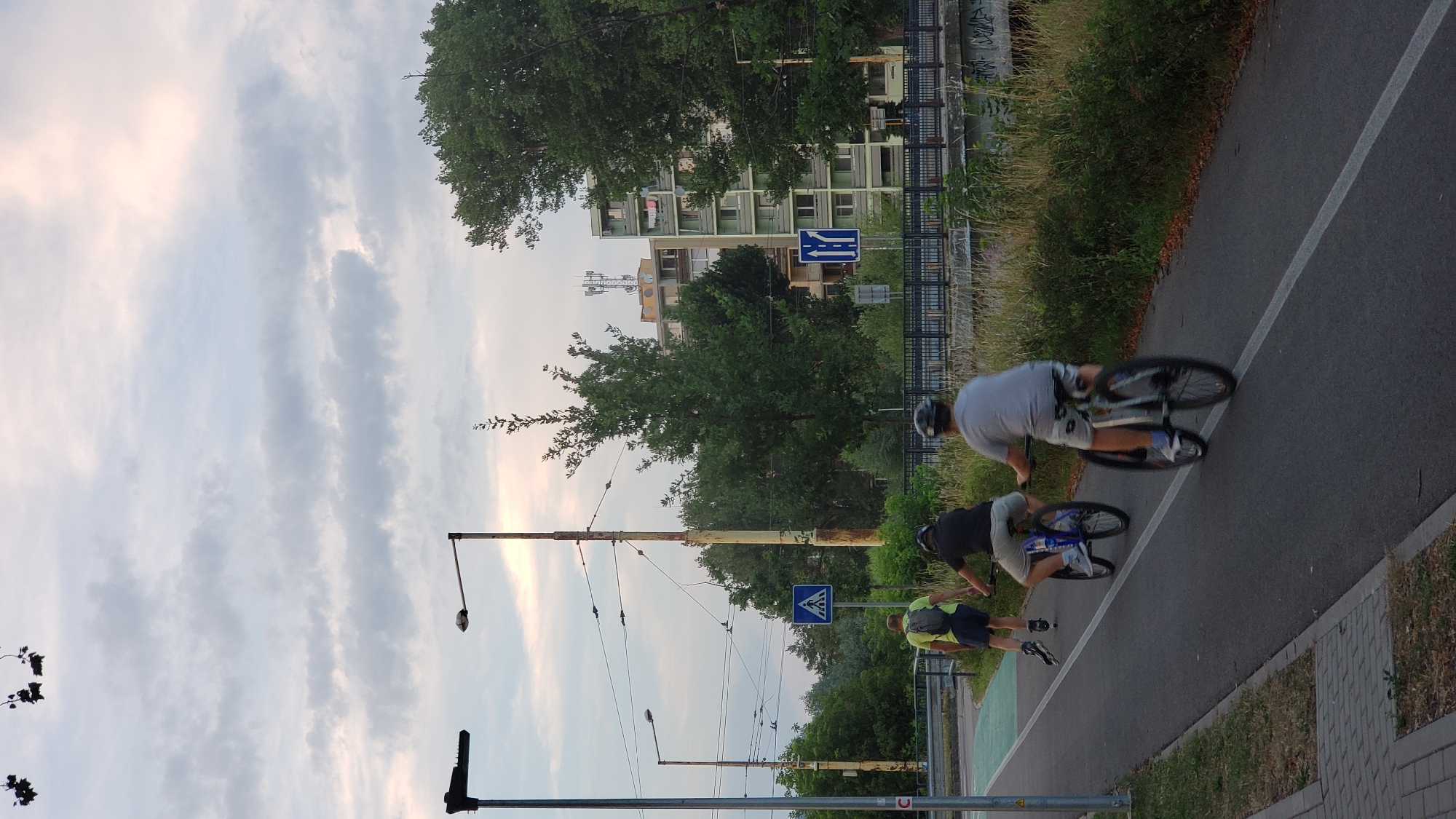 The image captures a view of a bike path in the city of Prešov, Slovakia. Three individuals are visible on the path. In the foreground, a person wearing a grey t-shirt and patterned shorts is riding a bicycle. Behind them, another person in a dark t-shirt and light shorts is also on a bicycle. To the left, a third individual is inline skating, wearing a bright yellow shirt and dark shorts. The bike path is paved and marked with white lines. The background features trees, buildings, streetlights, and utility poles. A pedestrian crossing sign and a traffic sign indicating lane guidance are also present. The sky above is partly cloudy with soft light. The overall scene suggests a recreational activity taking place in an urban environment.