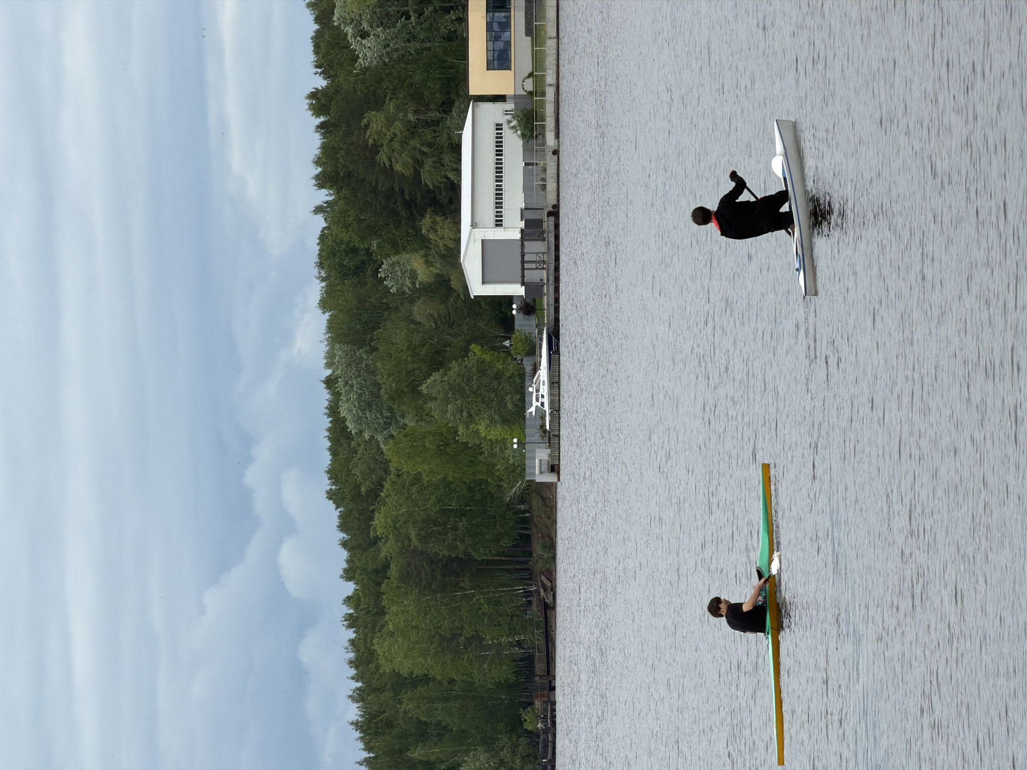 The image depicts a waterfront scene in Chaikovsky, Russia. Two individuals are rowing on the water. One is in a white kayak, wearing a black suit with a red collar and using a paddle. The other is in a single scull with yellow and green components, dressed in black.
The shoreline includes dense green trees and buildings, including a white industrial building with vertically ribbed panels and a beige structure on top. The sky is overcast with clouds.
