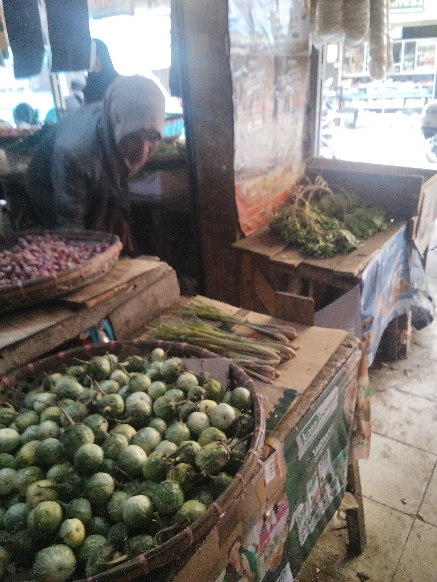 The image depicts an outdoor market stall in Cisaat, Indonesia. A woman, partially obscured and wearing a headscarf, is visible in the background, attending to her stall. The foreground is dominated by a woven basket filled with small, round green eggplants. To the left of the eggplants, a shallow woven basket holds a pile of red onions. Behind these, a wooden structure supports more produce, including leafy green vegetables. The stall is decorated with colorful advertising banners, some of which are partially visible. The ground is paved with tiles. The lighting suggests it is daytime, with bright light coming from above and behind the stall. The overall scene conveys a typical Indonesian market environment.
