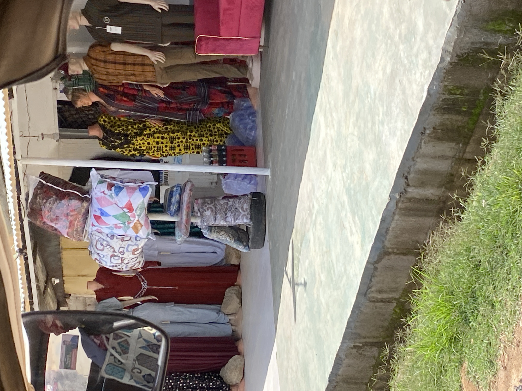 The image depicts an outdoor marketplace scene in Gura Top, Nigeria. The focus is on a stall filled with clothing displayed on mannequins and racks. Several mannequins are visible, dressed in various garments including patterned shirts, dresses, and trousers, suggesting a sale of apparel. The stall appears to be open-air, with a corrugated metal roof providing shade.
Objects in the stall include stacks of folded fabric, possibly textiles or additional clothing items, in different colors and patterns. One stack features a white fabric with colorful geometric designs. There are also some plastic bags, a red crate containing bottles, and what appears to be a spare tire. In the foreground, a car's side mirror reflects a poster with images and text, hinting at advertisement or information displays within the market.
The setting is a street or pathway, with a concrete surface and a raised drainage channel or curb on the right side, overgrown with grass. The lighting suggests it is daytime, with sunlight casting shadows, including a distinct shadow of a crossing or structure on the concrete surface. The overall atmosphere is that of a bustling local market, likely a daily trading activity. There is no indication of specific emotions or weather conditions beyond daylight. No legible text is visible on the clothing or main stall items, although the reflection in the mirror might contain some.