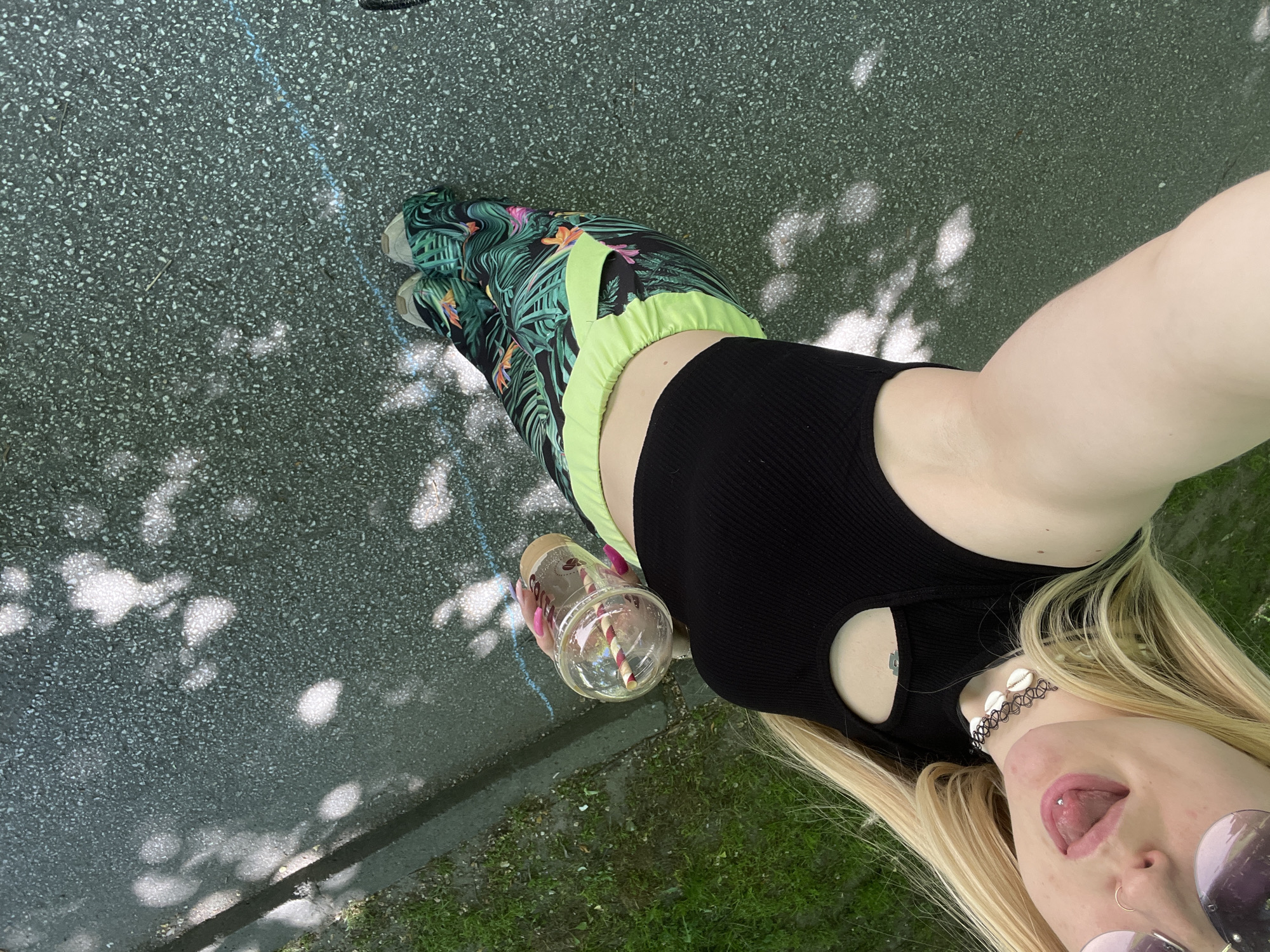 The image shows a person taking a selfie outdoors. The person is wearing a black crop top, tropical print pants with a neon green waistband, and sneakers with a similar print. They are holding a plastic cup with a straw. The location appears to be a paved area, possibly a sidewalk, with shadows cast by trees. The presence of grass at the edge of the pavement indicates a park or green space setting. There is no direct evidence in the image to confirm the location as Plovdiv, Bulgaria, but the details suggest a similar urban park environment. The person has long blonde hair, sunglasses, a choker necklace and a tongue piercing.