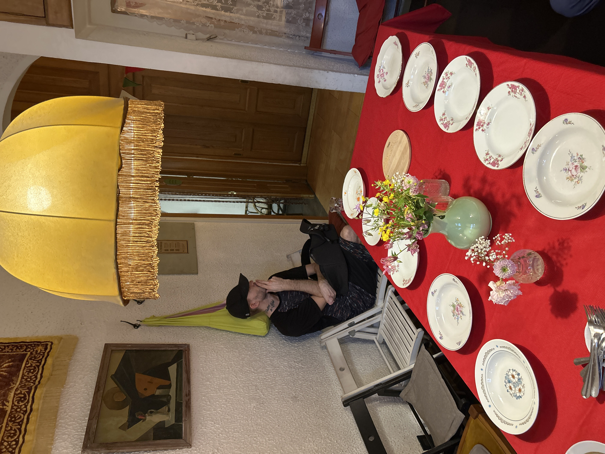 A man is sitting in a white chair in a room with white, textured walls. He is wearing a black baseball cap, a black t-shirt with a patterned design, and black pants. He is curled up in the chair, facing away from a red table set with floral-patterned plates, cutlery, and vases of flowers. A large yellow lampshade hangs overhead. The table is set for a meal. An abstract painting hangs on the wall next to the chair. The location appears to be inside a home or dining establishment. The specific city is not evident from the image.