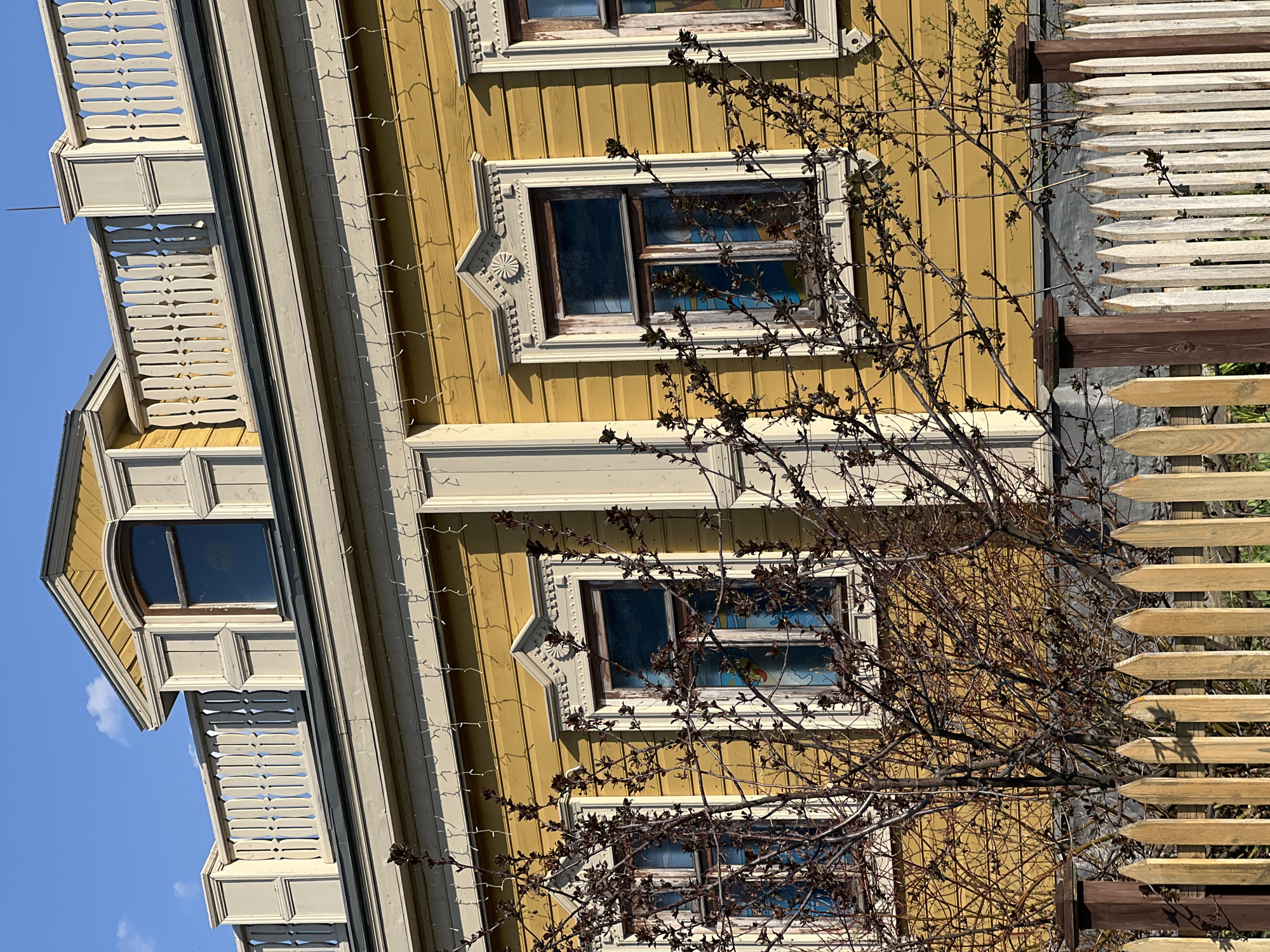 A yellow, three-story building with white trim and a white picket fence stands in Sviyazhsk, Russia. The building features three windows with ornate trim on the visible side and a balcony on the upper floors. A bare tree with spindly branches partially obscures the building and fence. The sky is blue.