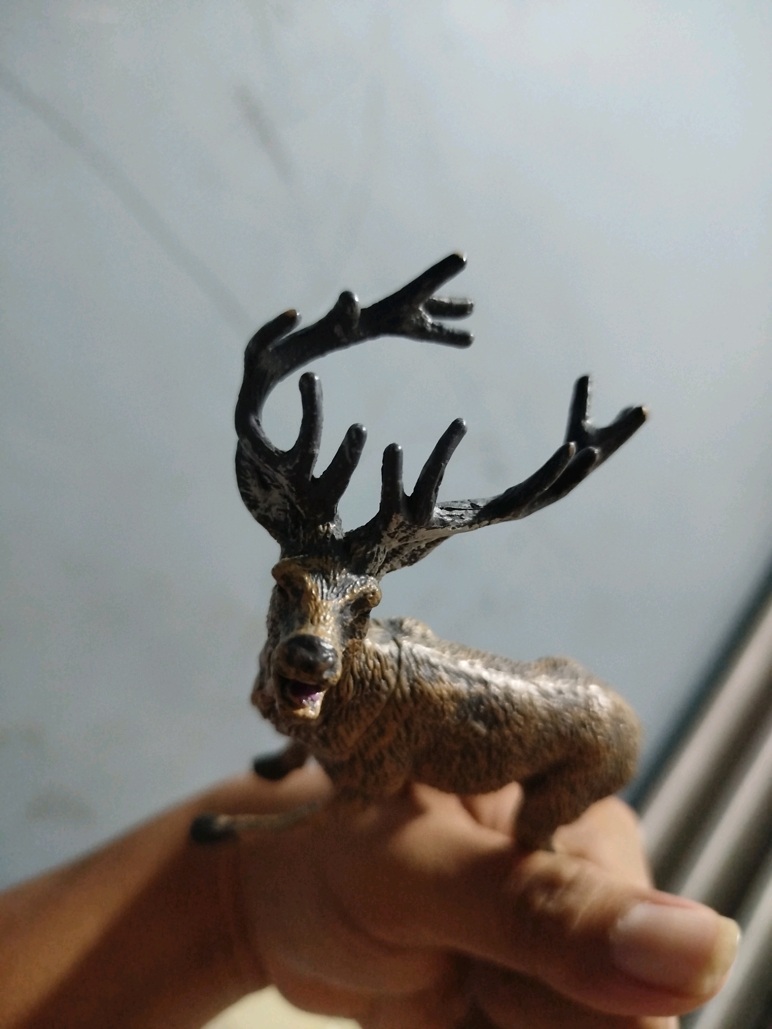 A toy figurine of a deer with large antlers is held in a person's hand. The deer figurine is brown with intricate detailing on its fur and antlers. The person's hand is visible in the foreground, with fingers curled around the figurine. The background is a light gray wall, possibly in a room in Mojoagung, Indonesia. The lighting suggests it is daytime, with shadows cast on the wall. The overall scene is a close-up shot of the figurine and the hand holding it.