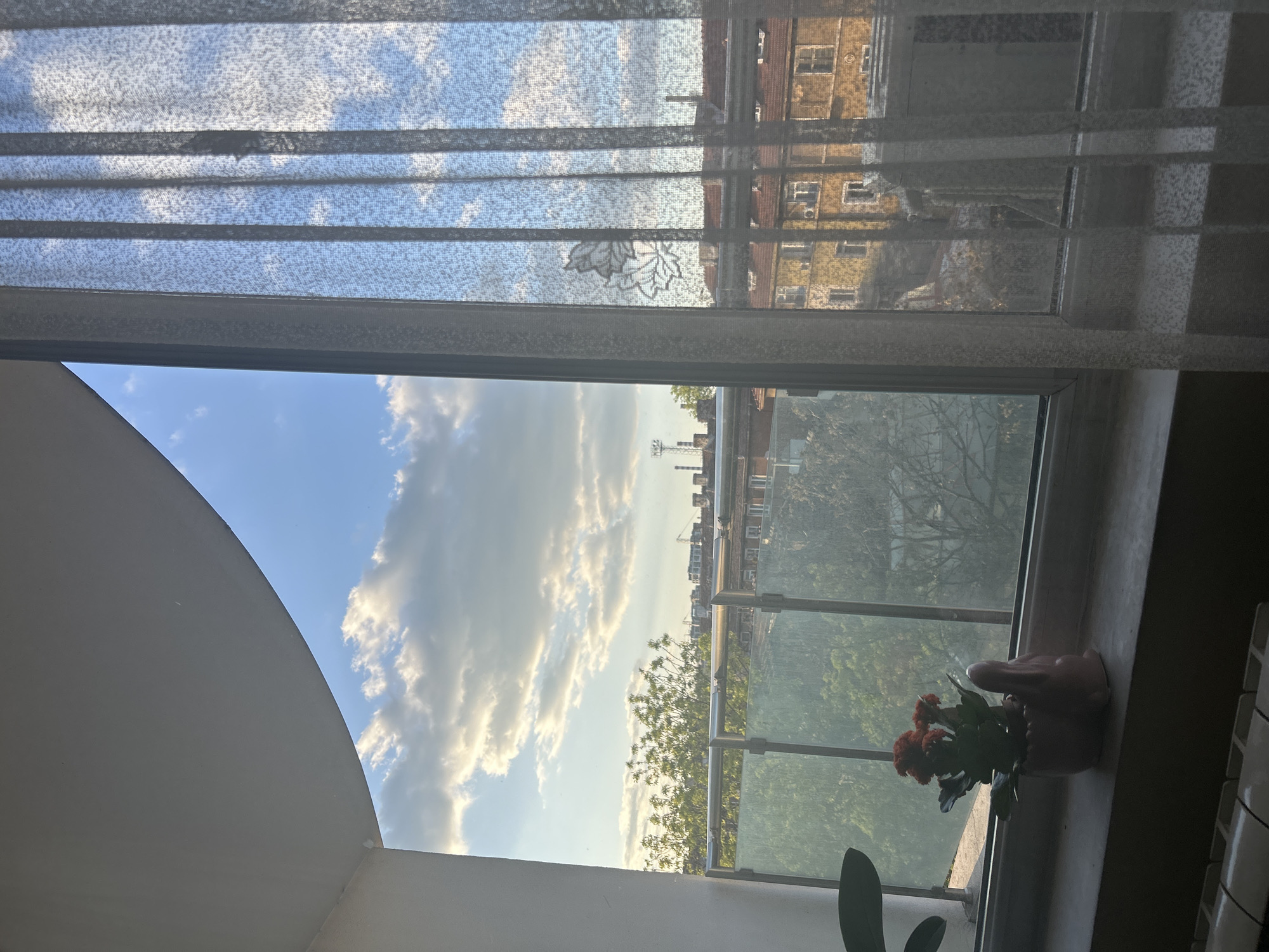 The image shows a view from a window. The upper part of the window is covered with semi-transparent blinds. Through the blinds and the open part of the window, one can see a building across the street, a blue sky with white clouds, and the cityscape, including a communications tower. A balcony with a glass railing is visible, as is a potted plant with red flowers sitting on the windowsill. The image was taken in Sofia, Bulgaria.