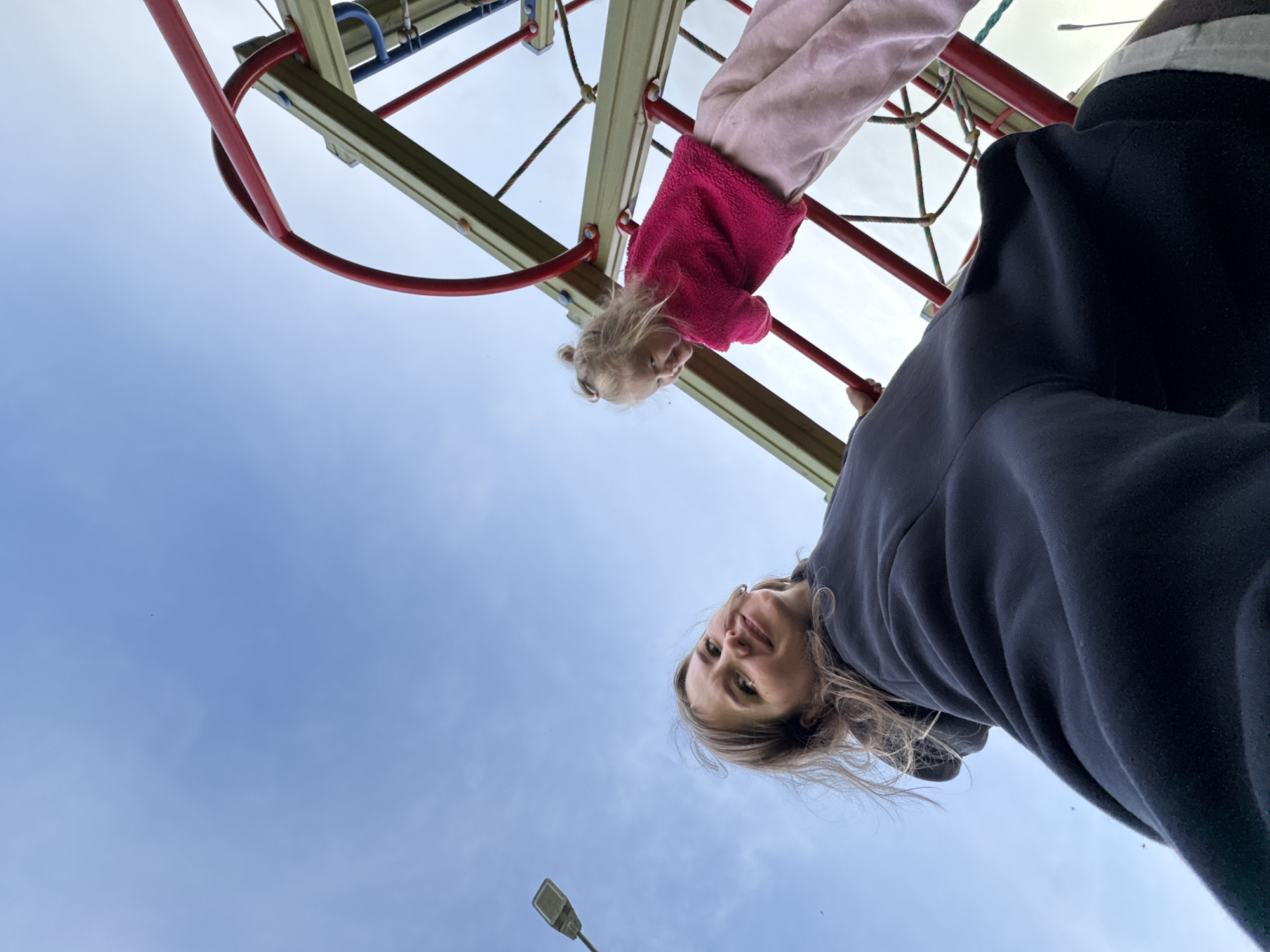 A low-angle selfie photograph captures an adult female and a young child outdoors in Горный, Russia. The adult female, positioned in the lower left foreground, has light brown hair and wears a dark-colored hooded sweatshirt. She looks upward towards the camera with a subtle smile. Her right arm, extending towards the lens, indicates she is holding the camera. In the upper right mid-ground, a young girl with blonde hair styled in a top knot is actively climbing a playground structure. She wears a bright pink long-sleeved fluffy top and light pink sweatpants, and displays a slight grin while looking towards the camera's general direction. The playground equipment consists of olive-green support beams, red metal bars, and rope elements. A grey streetlamp pole is visible against the light blue sky in the far left background. The sky is predominantly clear with faint, wispy clouds.