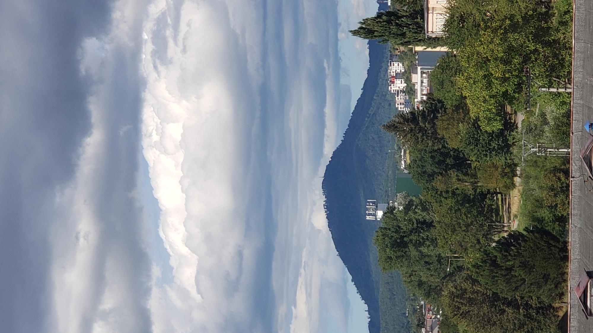 This image captures a panoramic view of the city of Prešov, Slovakia, dominated by the prominent peak of Šariš Castle (Šarišský hrad) in the distance. The castle hill, with its sparse vegetation, is silhouetted against a sky filled with a mix of dark storm clouds and bright white cumulus clouds. A small antenna is visible at the very summit of the hill.
In the mid-ground, a dense forest covers the lower slopes of the castle hill and extends towards the city. The foreground features a cluster of trees, with various shades of green foliage. Interspersed among the trees are modern buildings, including multi-story apartment complexes with white facades and red accents, as well as industrial structures like a tall white communication tower with a lattice design. The lower part of the image shows a glimpse of rooftops, suggesting a residential or urban area. The overall impression is a transition from natural landscape to urban development.