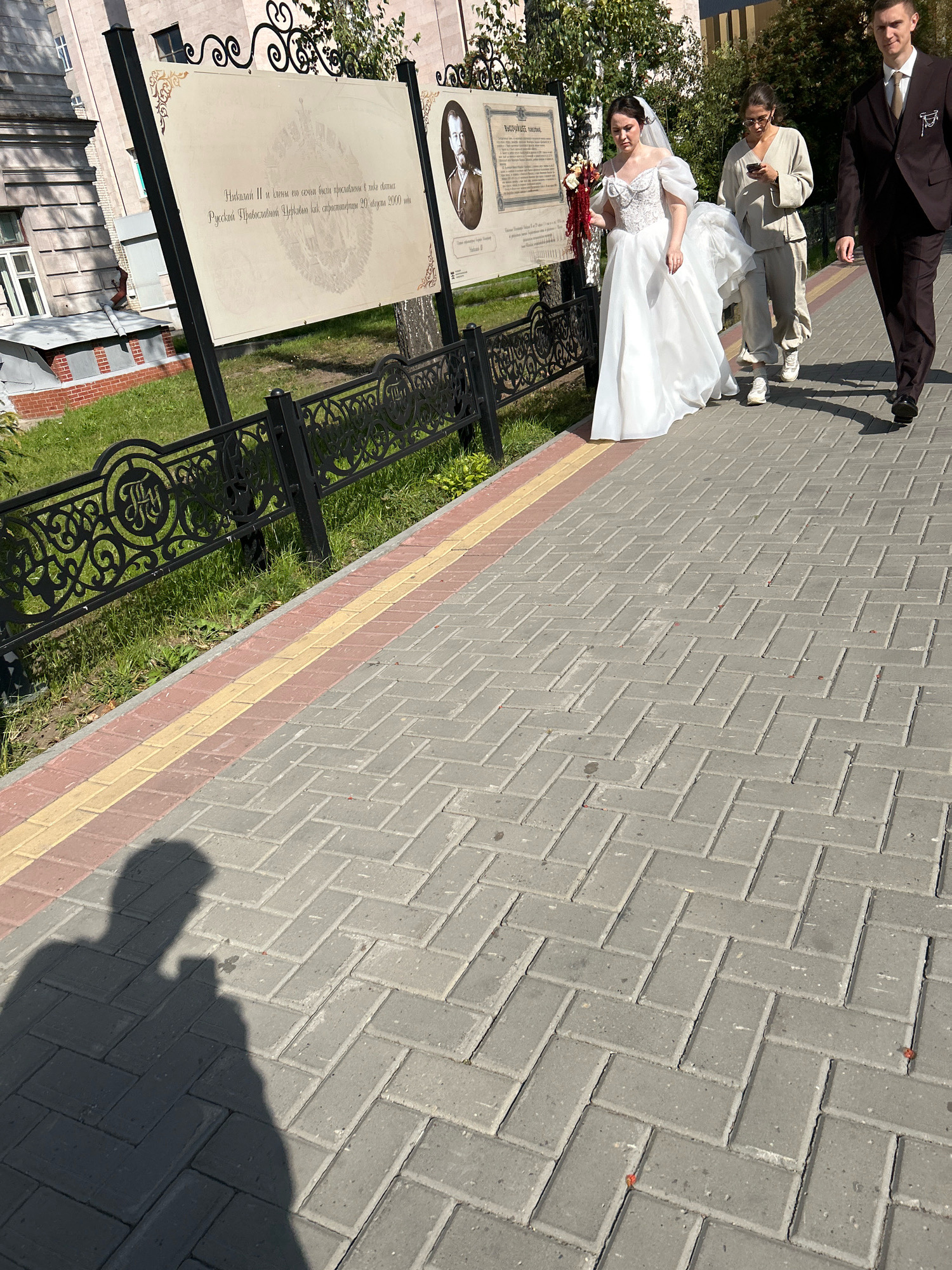 A bride in a white wedding dress, holding a bouquet, walks with two other individuals along a paved walkway in Tomsk, Russia. The bride is accompanied by a man in a dark suit and tie and a woman in casual light-colored clothing who is looking at her phone. Behind them, an information board details historical information, likely related to the city of Tomsk, as indicated by the text "Русской Православной Церкви, как страстотерпцы 20 августа 2000 года" which translates to "of the Russian Orthodox Church, as Passion-Bearers on August 20, 2000." The scene takes place outdoors on a sunny day, with a shadow of the photographer visible in the lower left of the image. The walkway is bordered by a lawn and a decorative metal fence.
