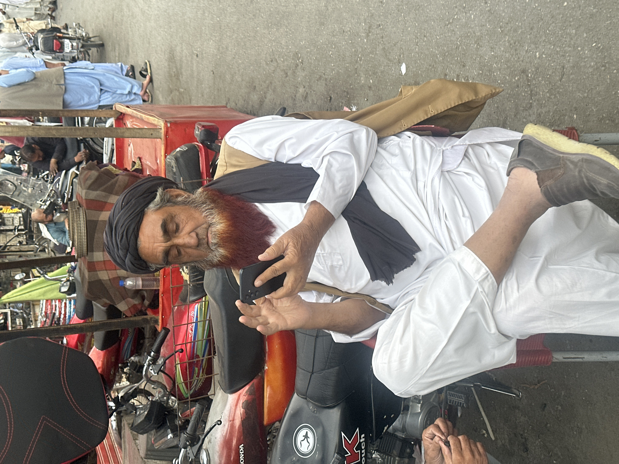 An elderly man with a long, reddish-orange beard and mustache, wearing a black turban, a white traditional long-sleeved tunic and trousers, a black scarf draped over his shoulders, and a brown vest, sits on a red metal stool. He holds a black smartphone in both hands, looking at its screen, with his left leg crossed over his right knee. He is wearing brown slip-on shoes with light-colored soles.
Behind and to the man's left are several motorcycles parked on an asphalt surface. Visible motorcycles include a black Honda CD 70 with a black seat and colorful graphic on the tank, a red Honda with a black seat, and another black motorcycle with a white bird logo and "KTOUCH" text. A pair of hands is visible in the lower left foreground, appearing to work on a small object. To the man's right, a red wooden stall or cart is partially visible, with a plastic water bottle placed on it.
In the midground and background, multiple men are present, some wearing light blue or white traditional attire. One man in a light blue tunic and beige vest is seen walking away from the viewer. Another man in a blue shirt leans over a motorcycle further left. The environment is an outdoor street or market setting, characterized by the presence of motorcycles and pedestrian activity on a grey asphalt ground. A green tarp or fabric is visible in the upper left background. The scene is located in the city of Unknown, Unknown.