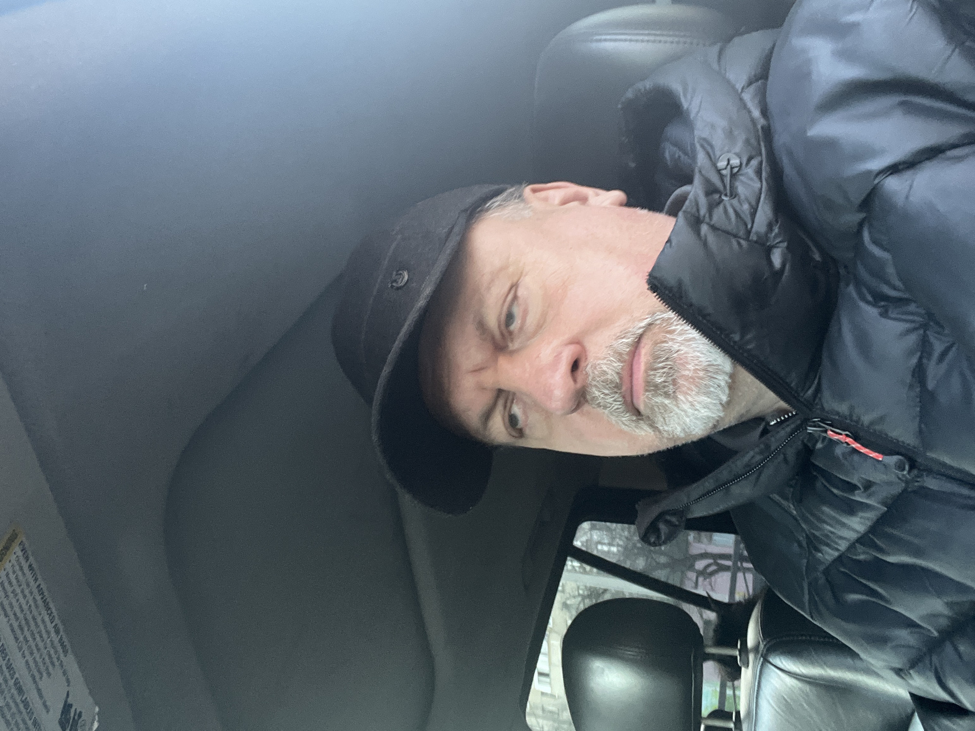 A man wearing a black hat and a black puffy jacket is sitting in what appears to be the driver's seat of a car. The interior of the car is visible, with the headrest of the passenger seat in the foreground and a warning label on the driver's side. Outside the car window, the blurry outlines of buildings suggest an urban environment, but it is impossible to pinpoint the location as Podolsk, Russia without further evidence. The man is looking directly at the camera. His face shows an older age with a white beard.