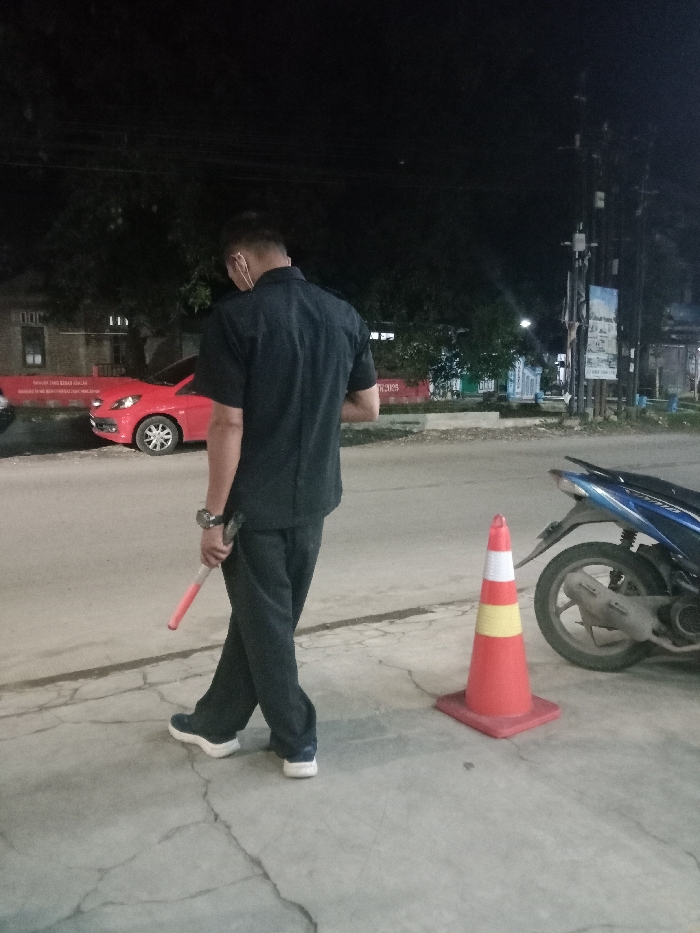 The image captures a nighttime street scene in Gladaksari, Indonesia. A male individual, seen from behind, walks from left to right across a concrete surface. He wears a dark short-sleeved collared shirt, dark trousers, and dark blue athletic shoes with white soles. A light-colored face mask hangs below his chin, and a watch is visible on his left wrist. He holds a red and white traffic baton in his right hand, angled downwards. To the individual's right, a red traffic cone with white and yellow bands stands on the concrete. Behind the cone, a blue and black scooter is parked, facing towards the left. In the left background, a red four-door sedan is parked on the roadside, with a red banner containing white text partially visible behind it. The environment features dark trees, utility poles with signs, and dimly lit buildings in the background, under ambient night lighting.