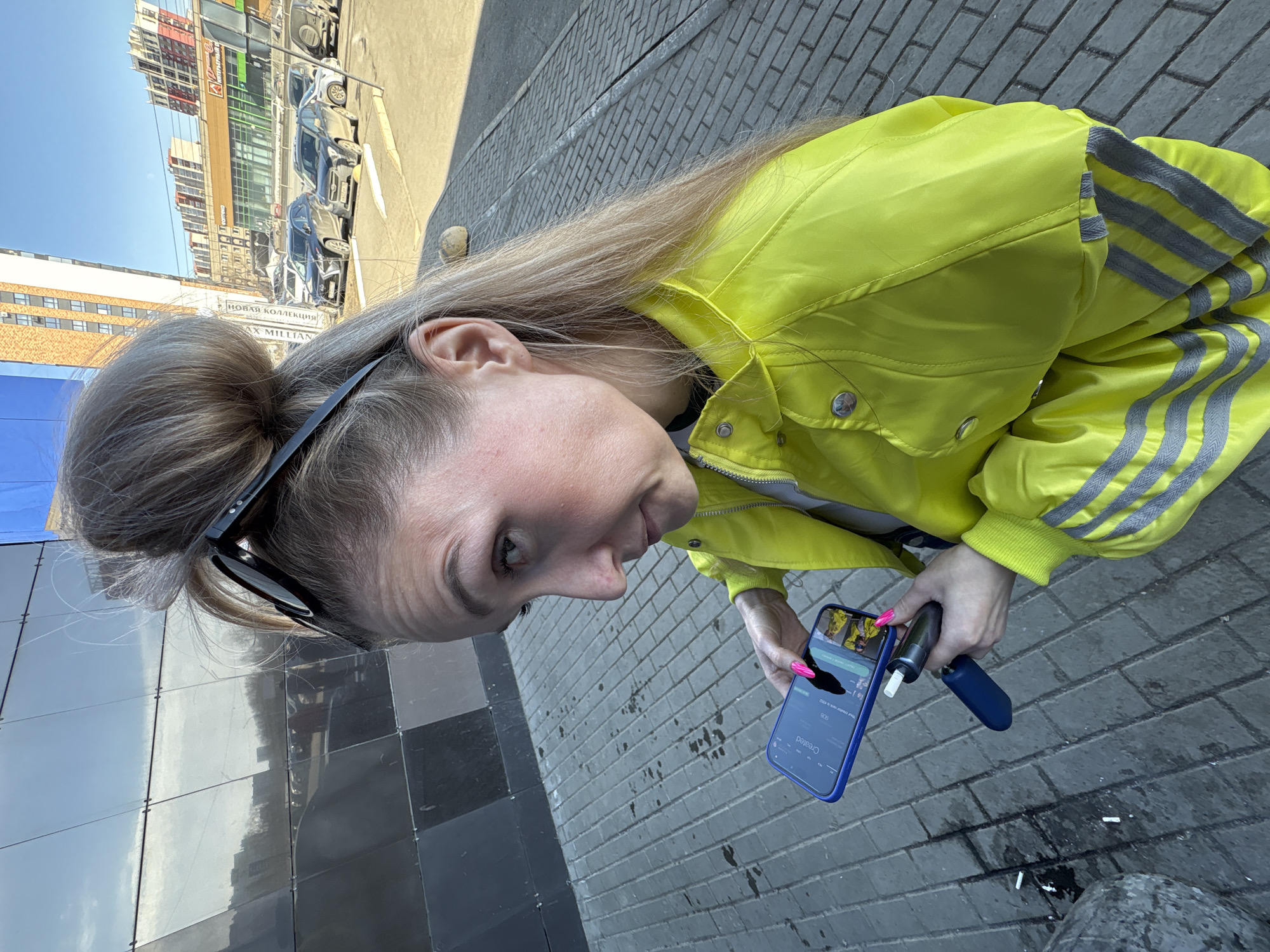 A close-up, high-angle shot depicts a female subject in an urban environment identified as Izhevsk, Russia, based on "18" region license plates visible on parked vehicles and Russian text ("ИНТЕРЬЕРНЫЙ САЛОН", "НОВАЯ КОЛЛЕКЦИЯ") on background buildings.
The subject, positioned in the foreground and center-right, is a blonde female with long hair, some of which is pulled into a bun atop her head, while other strands fall over her shoulders. Black sunglasses rest on top of her bun. She wears a bright, neon yellow jacket featuring grey striped reflective material on the sleeves and lower body, fastened with silver buttons and a zipper. Her fingernails are painted vivid pink. She directs her gaze slightly upwards and to the left, towards the camera.
In her left hand, she holds a blue-cased smartphone displaying an application with the word "Created" at the top and a small thumbnail image of a person in a yellow jacket. Her right hand holds a black and silver electronic cigarette (vape device) with a white filter-like tip, along with its detached blue bottom cap held beneath.
The immediate environment consists of grey rectangular brick paving. Several discarded white cigarette butts and dark liquid stains are present on the ground near the subject's feet. To her left, a modern building with a dark, reflective, segmented facade is visible, reflecting a cloudy sky and distant structures. The background, particularly to her right, reveals a street with multiple parked cars. Beyond the cars, several multi-story buildings are visible under a clear blue sky, including one with red and white horizontal accents and a sign for an "Interior Salon."