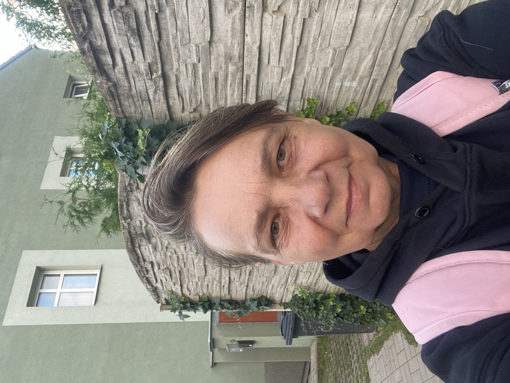 A woman is taking a selfie in front of a stone wall in Bílina, Czechia. She is wearing a black hoodie and a pink backpack. The wall is made of rough-hewn stones and is covered in ivy. Behind the wall, a pale green building with a white-framed window is visible. There are also some trees and bushes. To the left of the wall, there is a black trash can and a portion of a sidewalk made of light-colored pavers. The lighting suggests it is daytime.