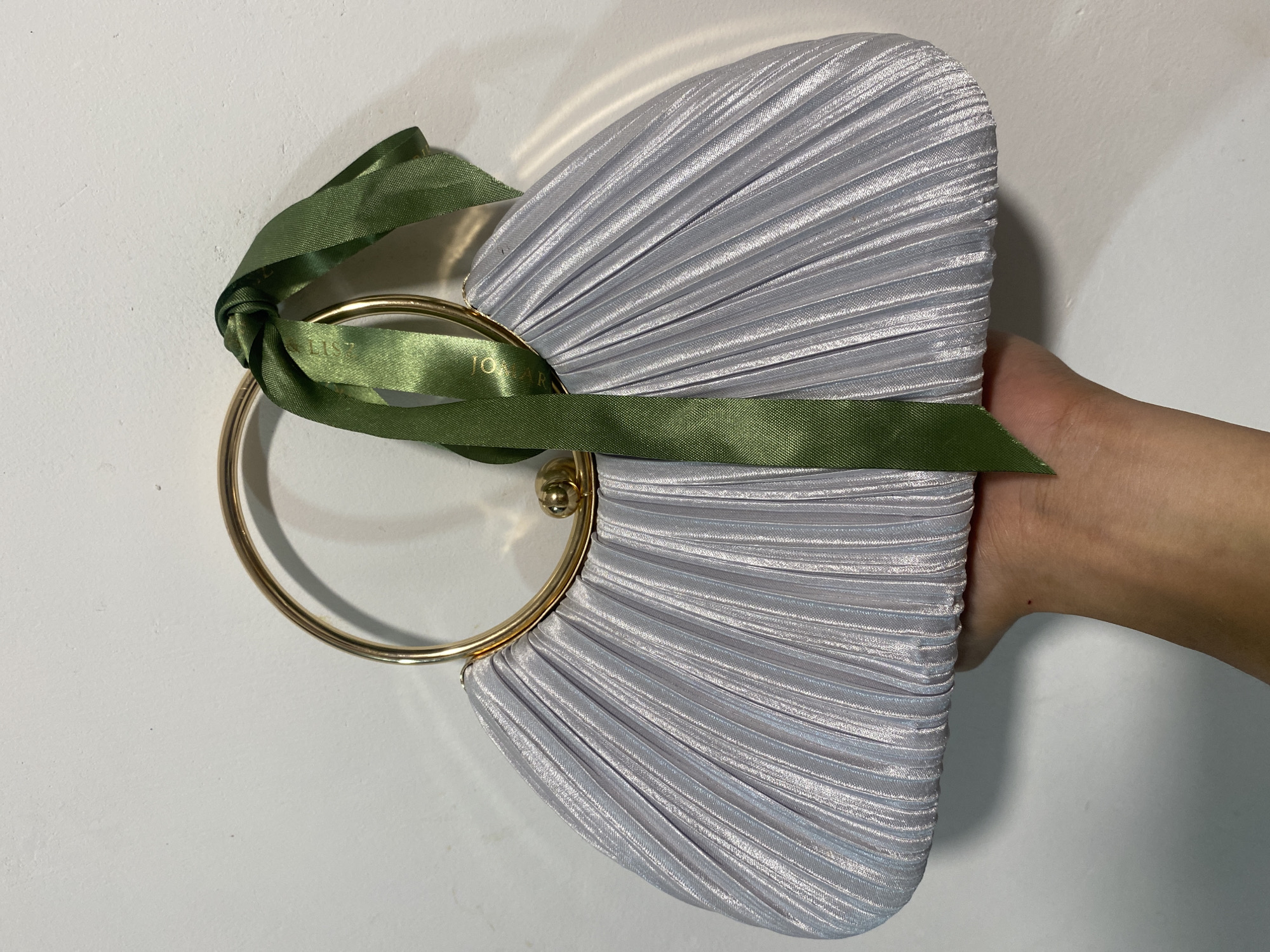 The image shows a hand holding a silver, pleated, fan-shaped clutch. The clutch has a gold, circular handle with a green ribbon tied around it. The background is a plain white surface. The location in Davao City, Philippines, is not identifiable from the image.