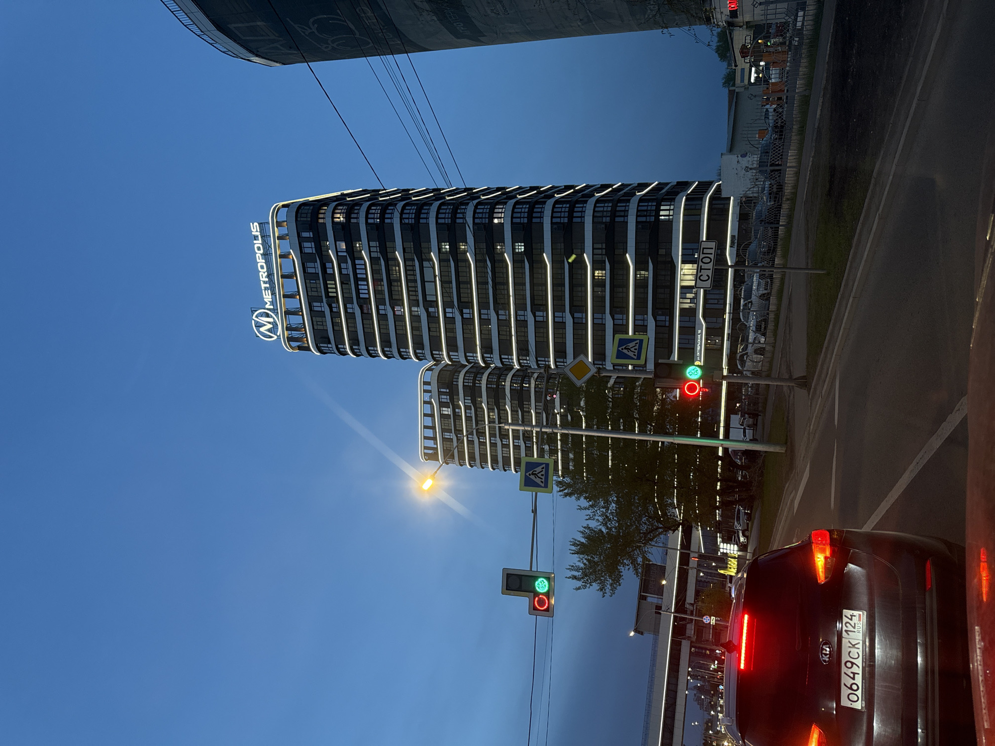 The image captures an urban scene in Krasnoyarsk, Russia, featuring the "Metropolis" residential complex. The complex consists of two modern high-rise buildings with white-trimmed balconies against a dark facade, illuminated by vertical light strips. The buildings are marked with the "Metropolis" logo. A black Kia sedan is visible in the foreground, its tail lights illuminated and license plate reading "O649CK 124 RUS" (likely indicating the Krasnoyarsk region). A traffic light pole displays a green light. Sidewalks and roadways are seen. The sky is a clear blue at dusk. Above the Metropolis towers is a building.