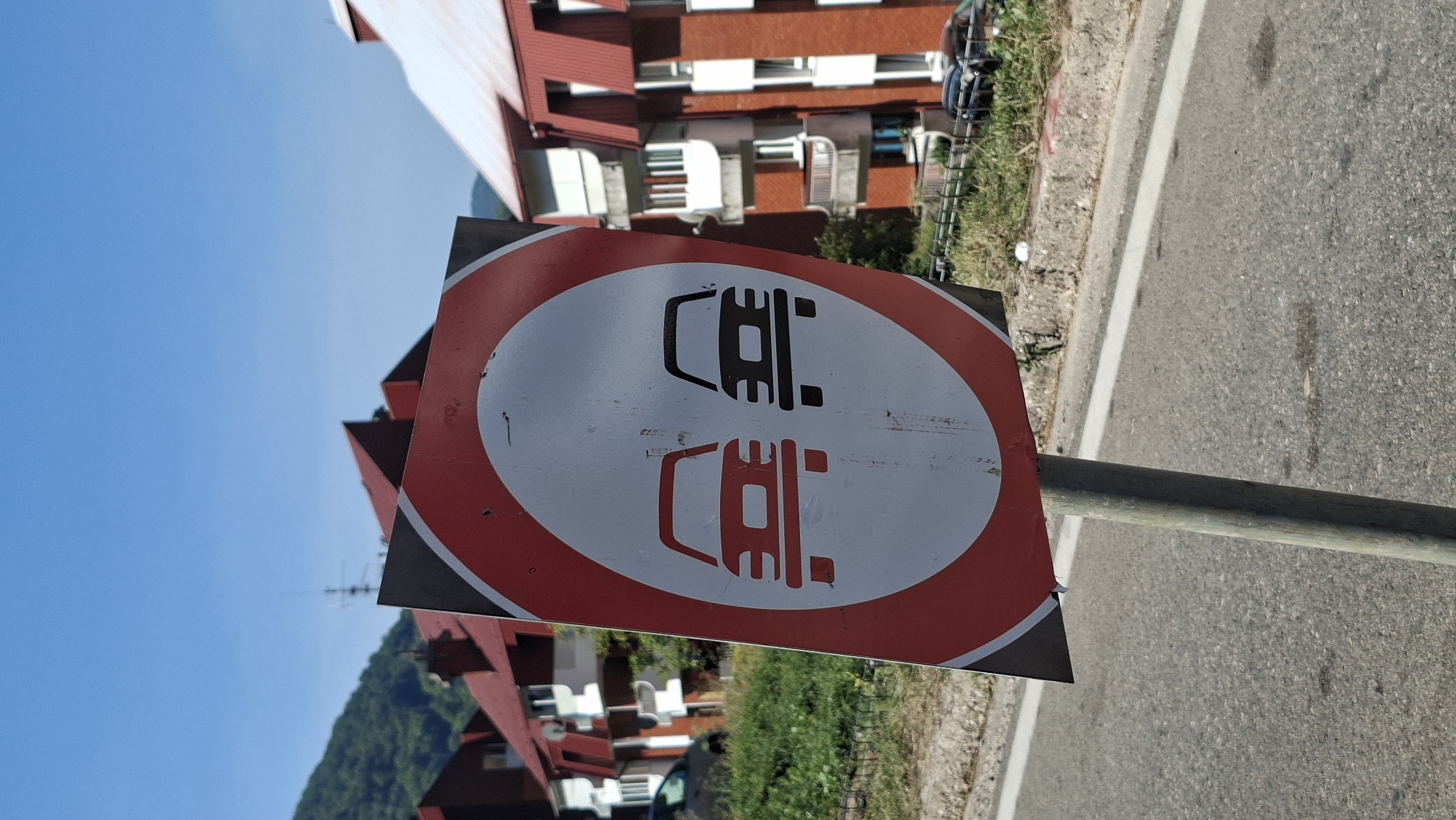 A tilted traffic sign, featuring a red circle with a white background, displays two car silhouettes within it: one red car on the left and a black car on the right. The sign is mounted on a metal pole in the foreground, positioned on the side of an asphalt road. In the background, red-roofed buildings with white accents are visible, nestled against a lush green hillside under a clear blue sky. The overall scene appears to be a road in or near the town of Plužine, Montenegro, characterized by a mountainous landscape and residential structures.