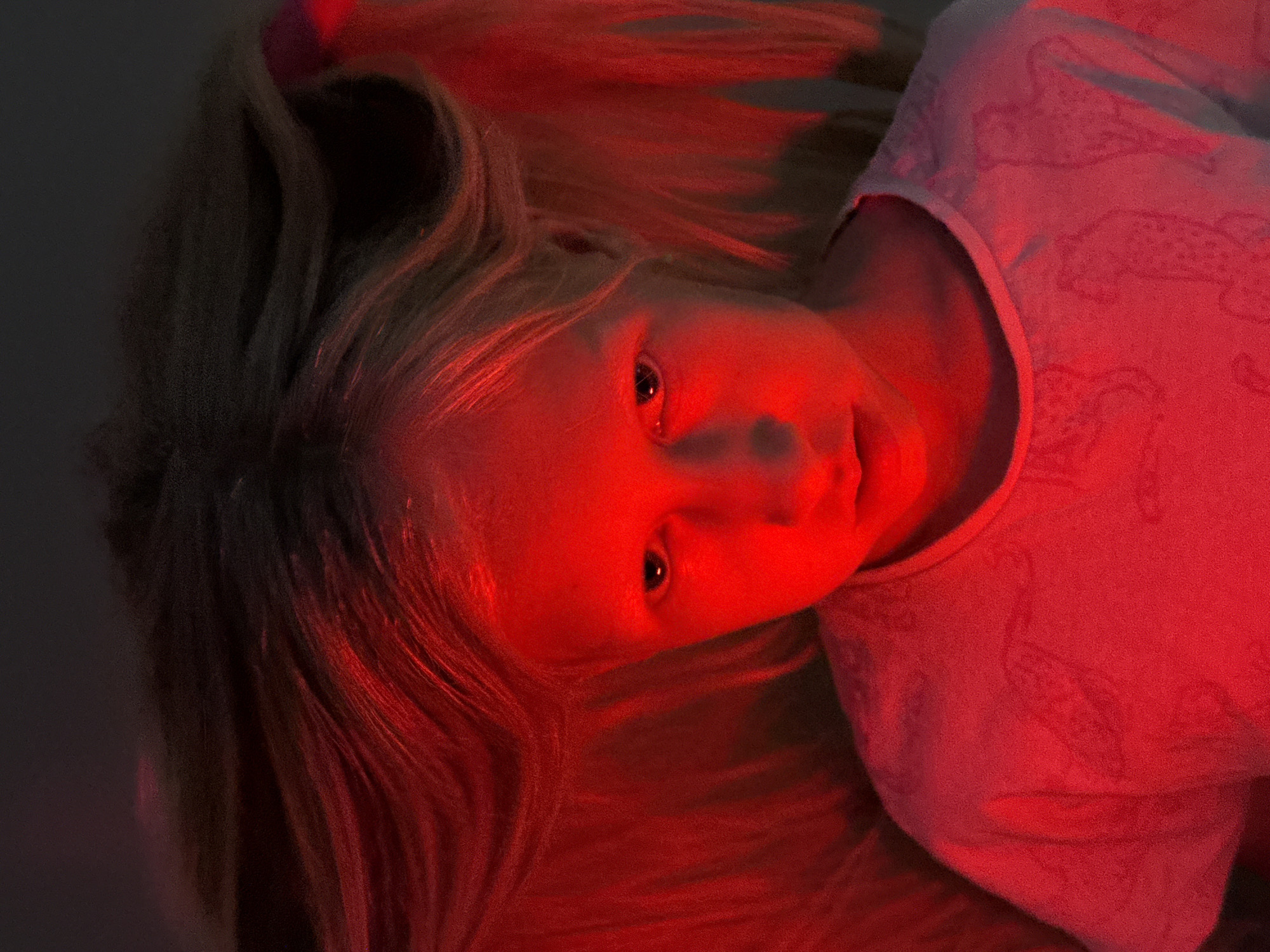 The image shows a young girl with blond hair, illuminated by red light. She is wearing a pink shirt with a patterned design. The background is mostly dark and out of focus. No information provided in the prompt allows to identify the location as Chaikovsky, Russia, or to make any other judgments about the scene.