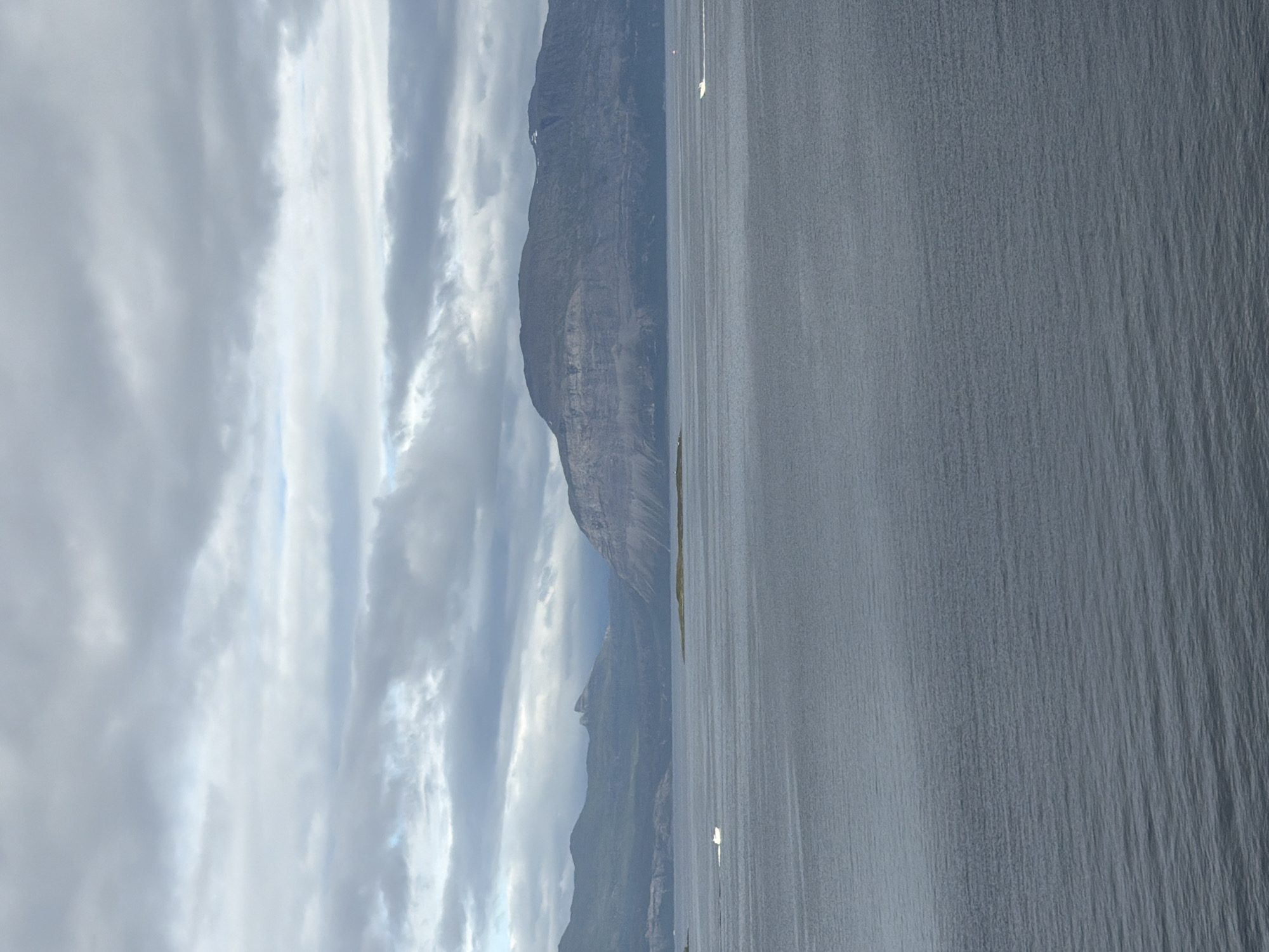 The image depicts a wide, calm body of water with a mountainous landscape in the background. The sky is overcast with dramatic, textured clouds. In the distance, a large, layered mountain range dominates the horizon, with some lighter patches possibly indicating snow. To the left, a smaller, jagged peak stands out against the softer hills. A small, vegetated island is visible in the middle ground. Two white boats are present on the water; one is closer to the left edge, and the other is further to the right, creating a wake. The water is a deep, muted blue-gray, showing gentle ripples. The scene is likely part of the Vågsfjorden, given its proximity to Harstad, Norway, a city known for its mountainous fjords and maritime activity. The overall impression is one of a serene, natural environment with human presence indicated by the boats.