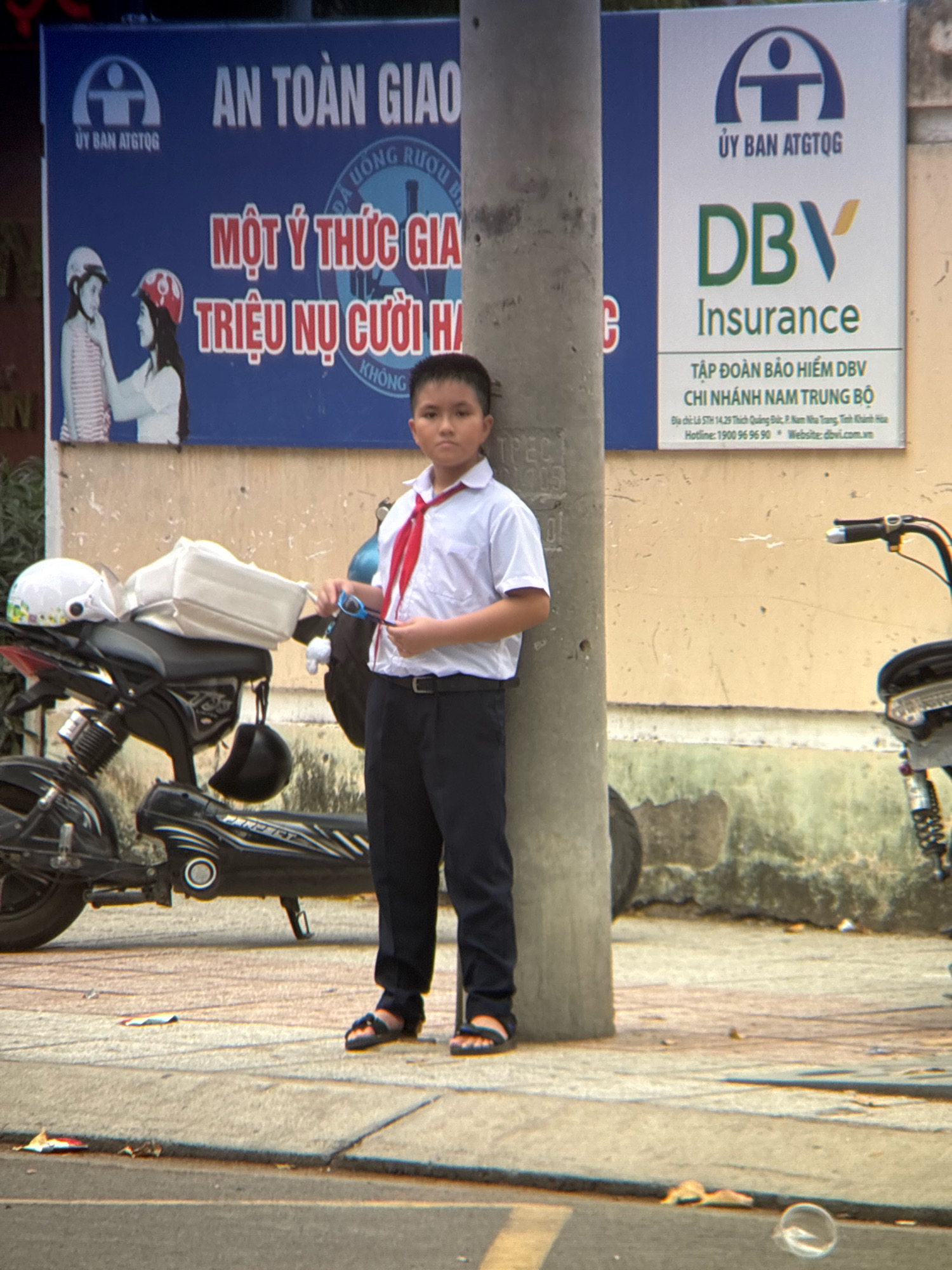 A young boy, likely a student given his uniform of a white collared shirt, red tie, and dark pants, stands next to a concrete utility pole on a sidewalk in Nha Trang, Vietnam. The boy is holding a pair of blue sunglasses in his right hand. Behind him, a black electric scooter is parked, equipped with two helmets and a white bag on its seat. The boy is positioned in front of a blue billboard that reads "AN TOAN GIAO" and "UY BAN ATGTQ" along with an image of two children wearing helmets. To the boy's right, a white billboard advertises "DBV Insurance" with Vietnamese text below, including an address and hotline for the Nam Trung Bo branch. The background shows a textured, weathered wall and some debris on the ground.