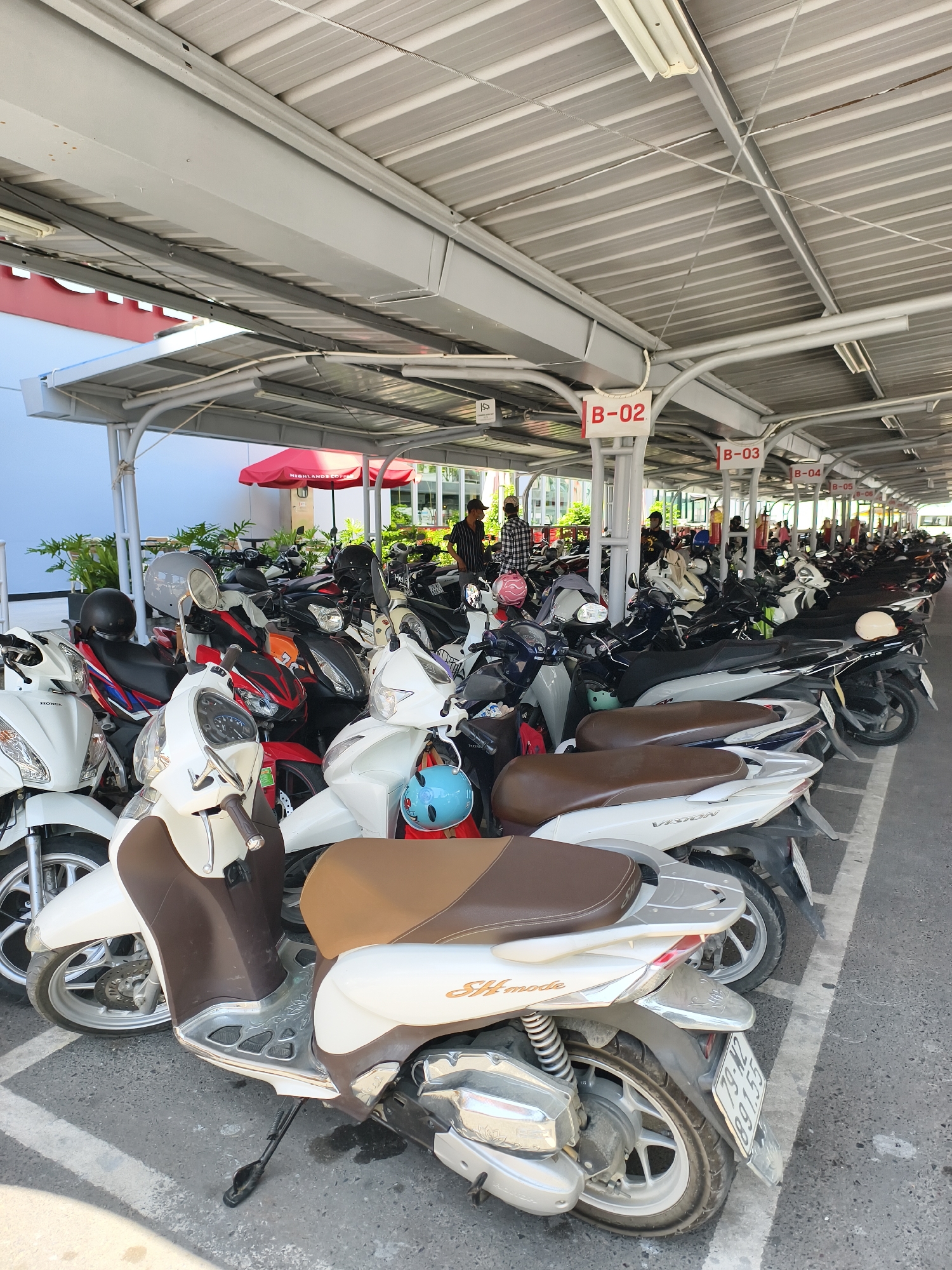 This image captures a crowded, covered parking area for scooters and motorcycles in Tay Nha Trang Ward, Nha Trang, Khánh Hòa Province, Vietnam. The location is confirmed by a visible license plate displaying "79-N2 891.55" (79 denoting Khánh Hòa Province) and a red "HIGHLANDS COFFEE" umbrella in the background. The parking structure consists of a grey corrugated metal roof, supported by grey beams and white pillars, with exposed electrical wiring and fluorescent light fixtures overhead. The ground is an asphalt or concrete surface, marked with faded white parking lines.
Numerous scooters and motorcycles of various models, including Honda SH mode and Honda Vision, are parked densely in rows, displaying colors predominantly white, black, and red, with some featuring brown seats. Multiple helmets in colors such as grey, black, pink, and blue are resting on the seats or handlebars of the parked vehicles. White signs with red text, such as "B-02", "B-03", "B-04", and "B-05", delineate parking sections. In the mid-ground and background, several individuals, dressed in casual attire, are seen standing or moving between the parked vehicles. Beyond the parking area, green foliage is visible, along with the aforementioned red umbrella, indicating the proximity of a commercial establishment.