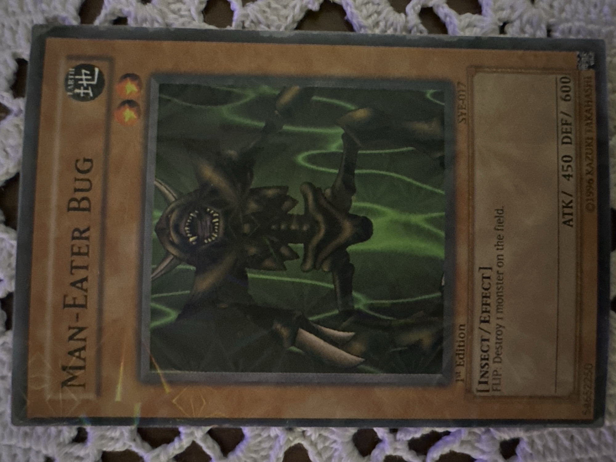 The image shows a "Man-Eater Bug" Yu-Gi-Oh! trading card. The card is in English and appears to be a common card. The card is tan/brown with a picture of a green insect in the center. The card's type is EARTH, its level is 2, and its ATK/DEF are 450/600. The text on the card indicates it is an insect/effect monster with the effect, "FLIP: Destroy 1 monster on the field." The card is surrounded by a white crocheted material. The image contains no indication of being located in the city of Sofia, Bulgaria.