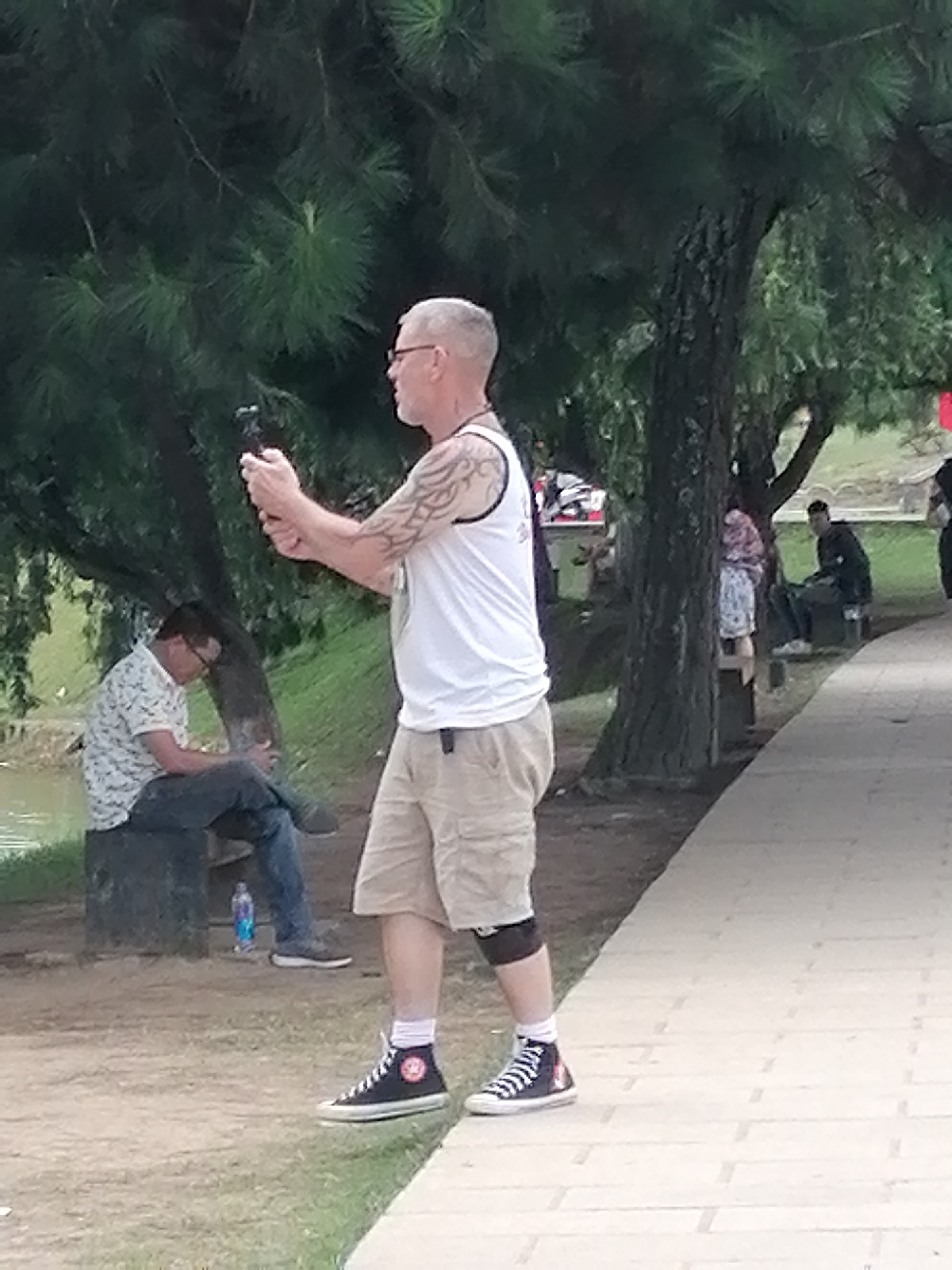 The image captures a street scene in Da Lat, Vietnam. In the foreground, a man with a grey beard and glasses is taking a picture with a smartphone mounted on a gimbal. He is wearing a white tank top with a black trim, khaki shorts, black high-top sneakers with white laces, and a black knee brace. He has tattoos on his left arm. To the left of the foreground man, another man is sitting on a dark grey rock. He is wearing a white collared shirt with a floral pattern and blue jeans. A blue plastic bottle is on the ground next to him. Behind them, a large tree with dark bark stands prominently, with lush green foliage surrounding it. Further in the background, other individuals are visible, including a woman wearing a patterned dress and a man in a dark jacket. A paved walkway runs through the scene, with grass and dirt areas adjacent to it.