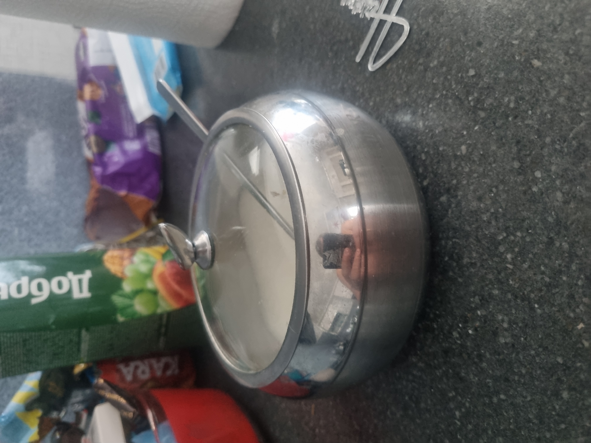 A stainless steel sugar bowl with a glass lid sits on a dark gray speckled countertop. The sugar bowl is filled with white sugar, and a metal spoon rests on top of the lid. In the background, a green juice carton with the Russian word "Добрый" (Dobry) is visible, along with a red container and a package of purple and white snacks. To the right of the sugar bowl, a white plastic object, possibly a hair clip, is on the counter. A reflection of a person holding a smartphone is visible on the side of the sugar bowl. The image appears to be taken indoors, possibly in a kitchen or pantry. There is no specific indication of the location being in the city of Samara, Russia.
