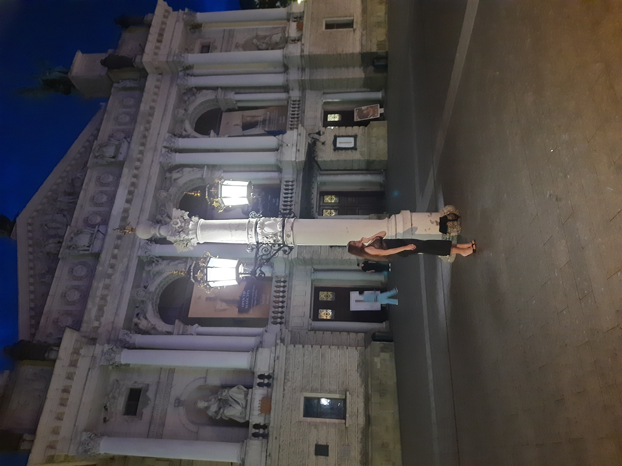 The image depicts the Lviv Opera House at night. A woman in a black dress stands near a lamppost in the foreground, looking to the right. The woman is wearing heels and carrying a bag. In the background, there are two figures walking, one in a light blue outfit and another in dark clothing. The Lviv Opera House is a grand building with classical architecture, featuring columns, ornate carvings, and large windows. The sky is dark blue, indicating it is nighttime.