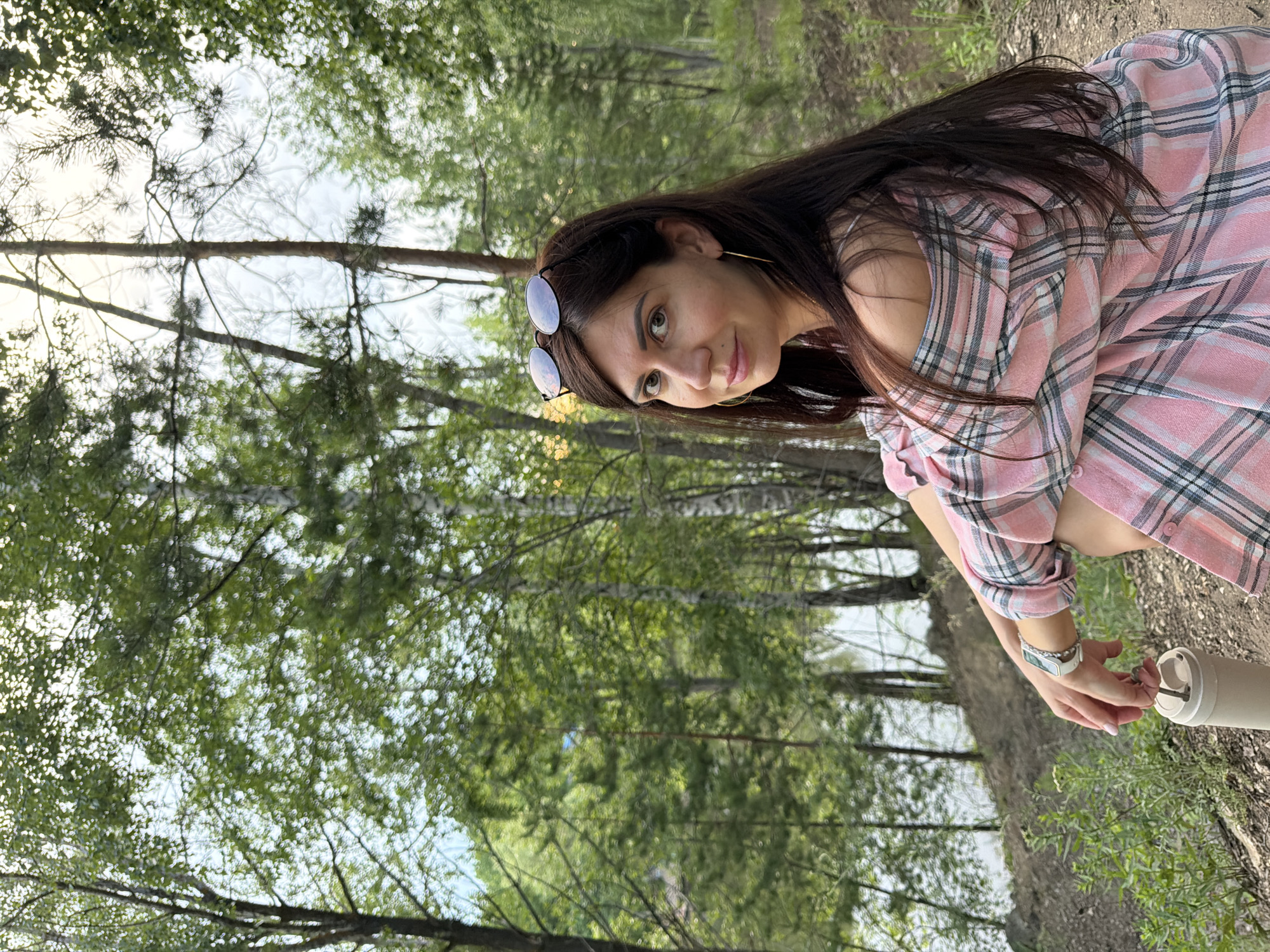 A woman with long, dark hair and sunglasses perched on her head is sitting on a dirt path in a forest near a body of water. She is wearing a pink and plaid off-the-shoulder shirt and a white watch. She is holding a white thermos and a key in her hand. The background features trees, foliage, and a glimpse of a lake. This scene is located in Volzhskiy, Russia, likely near the Volga River.