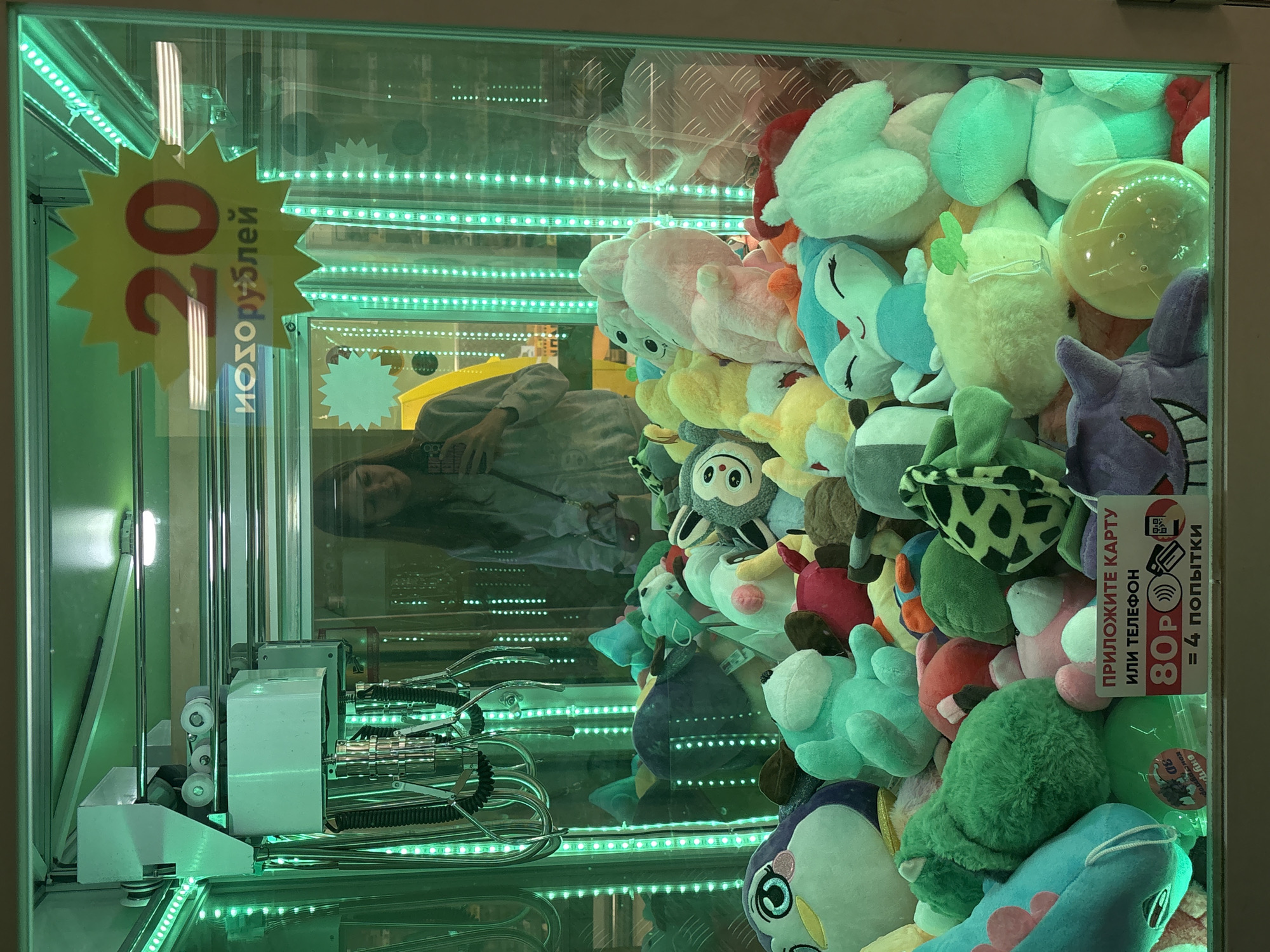 The image shows the inside of a claw machine filled with plush toys. A yellow starburst sign reads "20" and "Изорублей" (Rubles). A woman is reflected in the glass of the machine, wearing a gray sweatshirt. Another sign shows "80P = 4 попытки" (80 Rubles = 4 attempts).