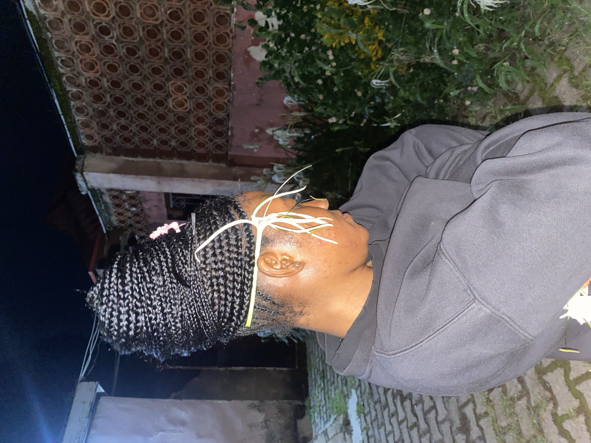 A young Black woman with braided hair is positioned outdoors at night, facing to the left and looking downwards. Her hair is styled into a large, intricate bun on top of her head, adorned with a small pink flower. Thin white flowers are also woven into her hair, some extending to frame her face. She is wearing a dark grey hooded sweatshirt and has her arms crossed. Her head is slightly bowed, and her eyes are not clearly visible.
The background suggests an urban or semi-urban environment in Jos, Nigeria. To her left, there is a building with a light-colored wall and what appears to be a paved walkway with interlocking bricks. To her right, a patterned brick wall, characteristic of some Jos architecture, runs horizontally. Beyond the brick wall, there is lush green foliage with scattered white and yellow flowers, indicating a garden or overgrown vegetation. The overall lighting suggests artificial illumination, consistent with nighttime. The scene appears to be a residential or public area within the city.