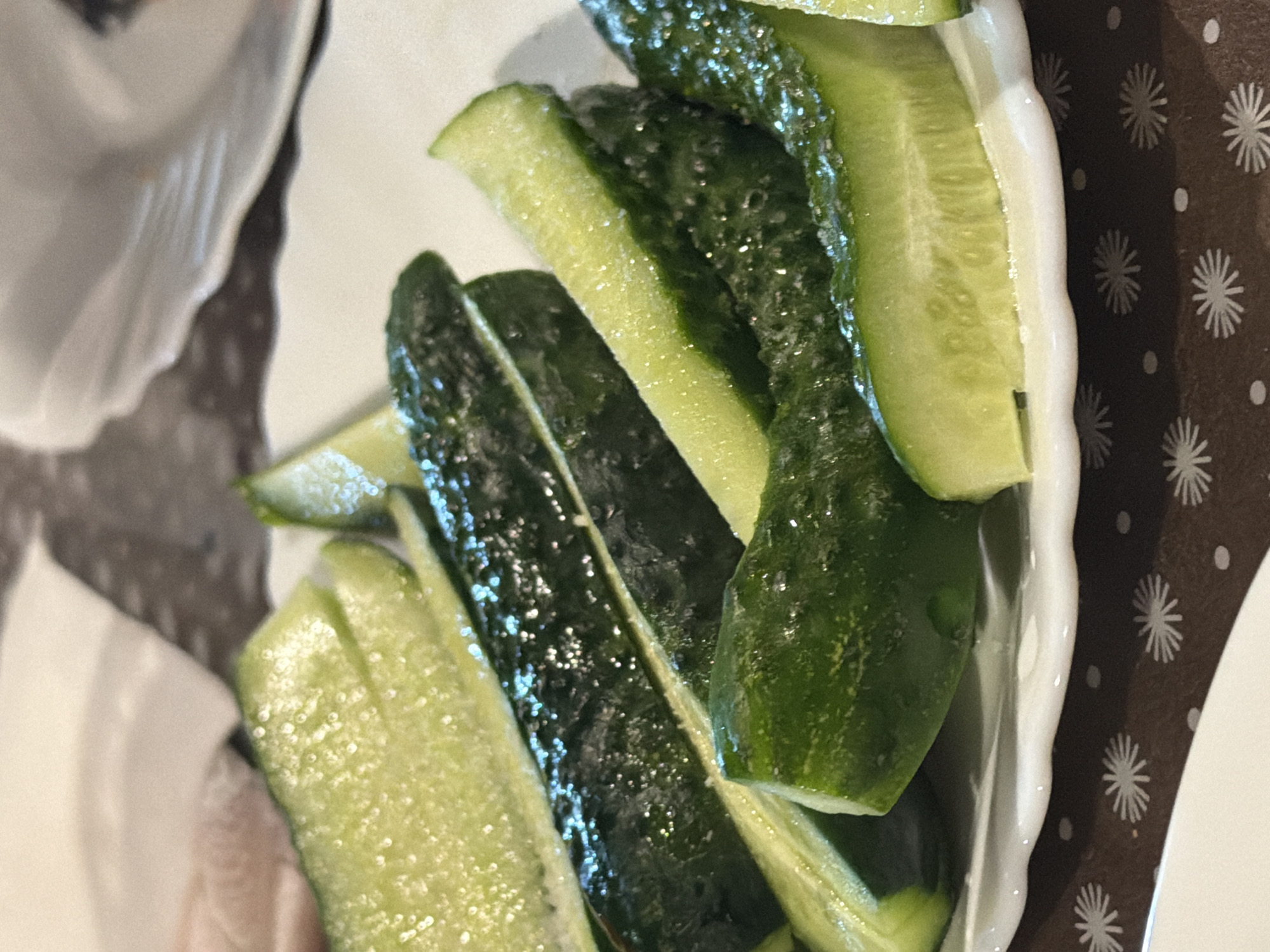 This is a close-up, eye-level, indoor shot of sliced cucumbers on a white, decorative plate. The plate is placed on a brown tablecloth with a pattern of white asterisks and dots. The cucumbers are sliced lengthwise and appear to be fresh, with visible seeds and skin. Some slices are glistening, suggesting they may be wet or seasoned. The lighting is bright, highlighting the textures of the food. There are no people or any identifiable landmarks of Sochi, Russia visible in the image.