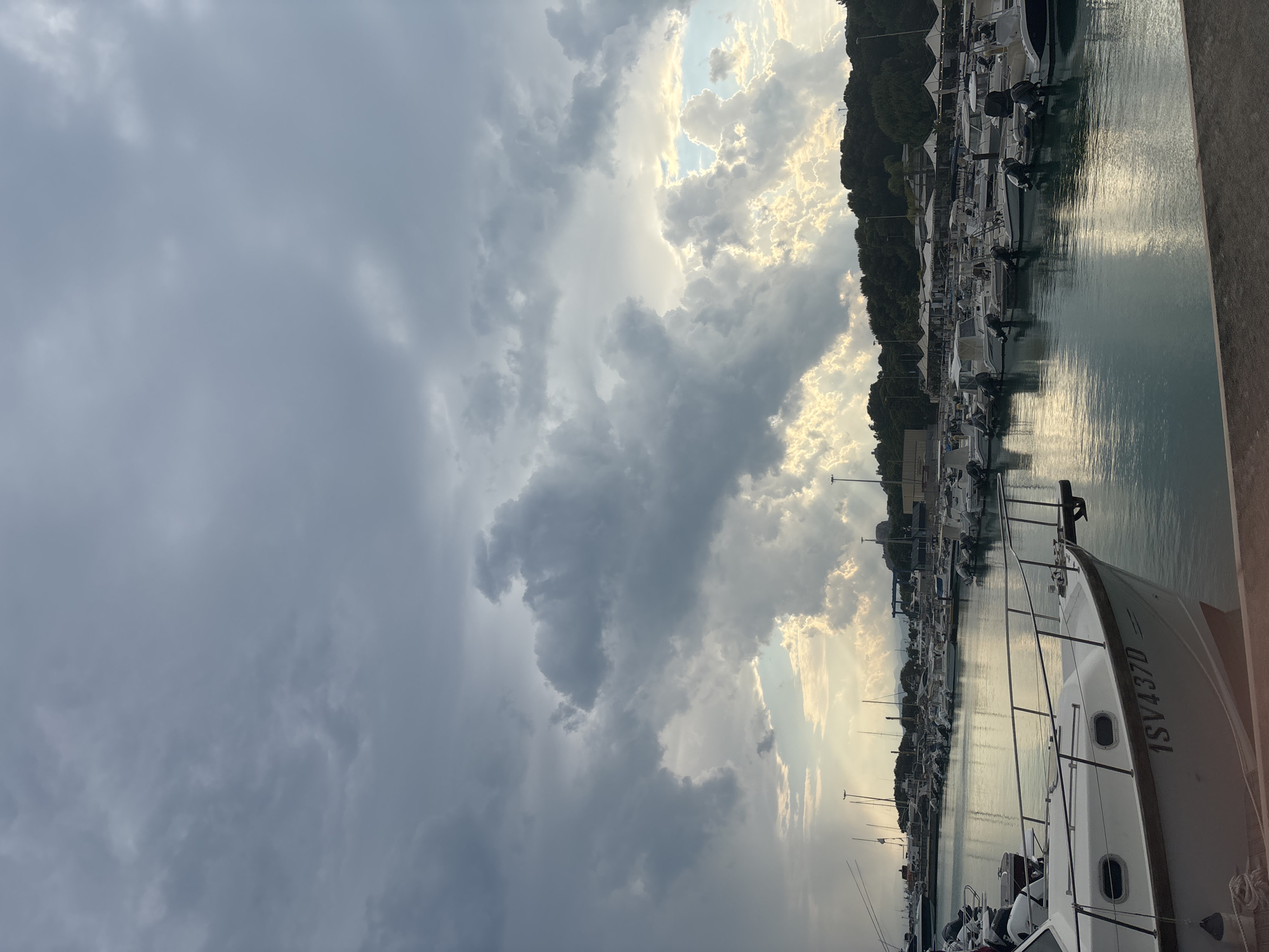 This media file depicts a marina at Roccella Ionica, Italy. The scene is dominated by a dramatic sky filled with both dark, heavy storm clouds and patches of bright sunlight breaking through, suggesting a dynamic weather event. The water is calm and reflects the sky and the line of docked boats. The boats, predominantly white with some dark accents, are moored in orderly rows. They appear to be a mix of motorboats and sailboats. Some of the boats have outboard motors. The hull of the closest boat in the foreground has the text "1SV437D" clearly visible.
Behind the marina, a dense line of trees forms a dark, verdant backdrop against the sky, with some buildings partially visible amongst the foliage. The time of day appears to be late afternoon or early evening, given the quality of the light. The overall mood is one of atmospheric tension due to the weather, contrasted with the peaceful stillness of the harbor. No people are clearly visible in the image.